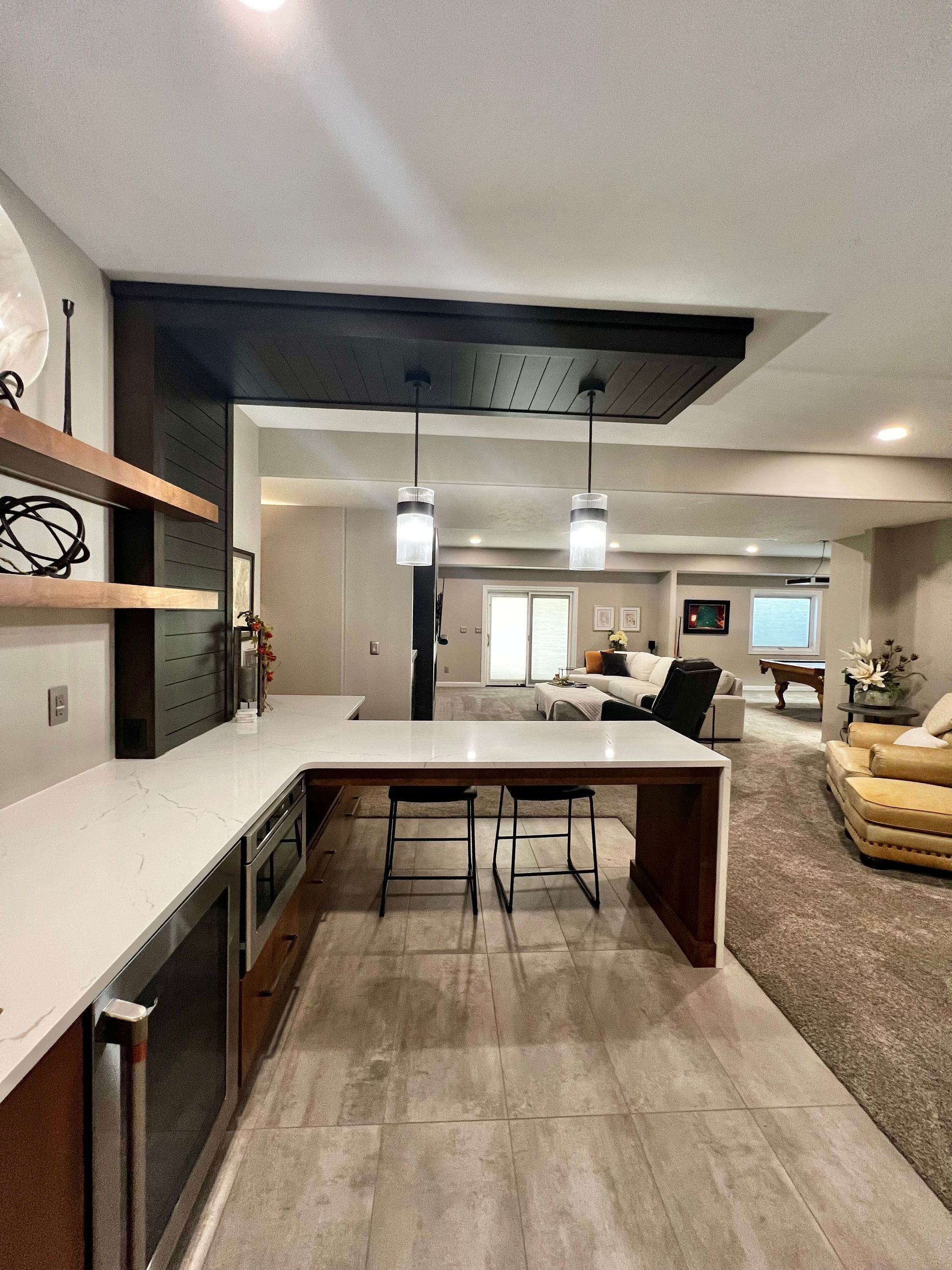 A modern home bar with white counters and dark wood trim, overlooking an open-plan living room with a neutral carpet.