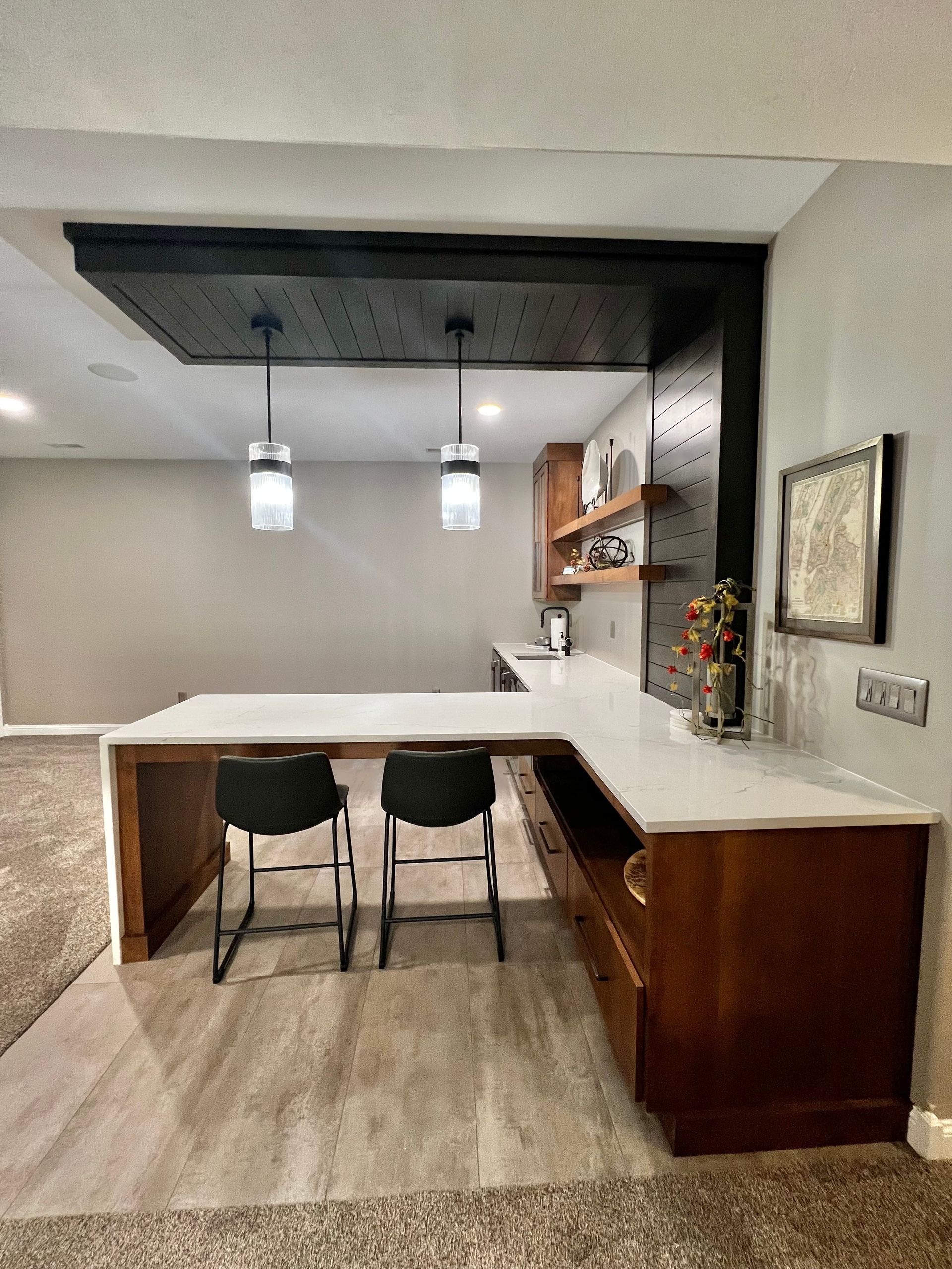 A kitchen peninsula with white countertops, two black bar stools, and a black pegboard ceiling feature with two pendants.