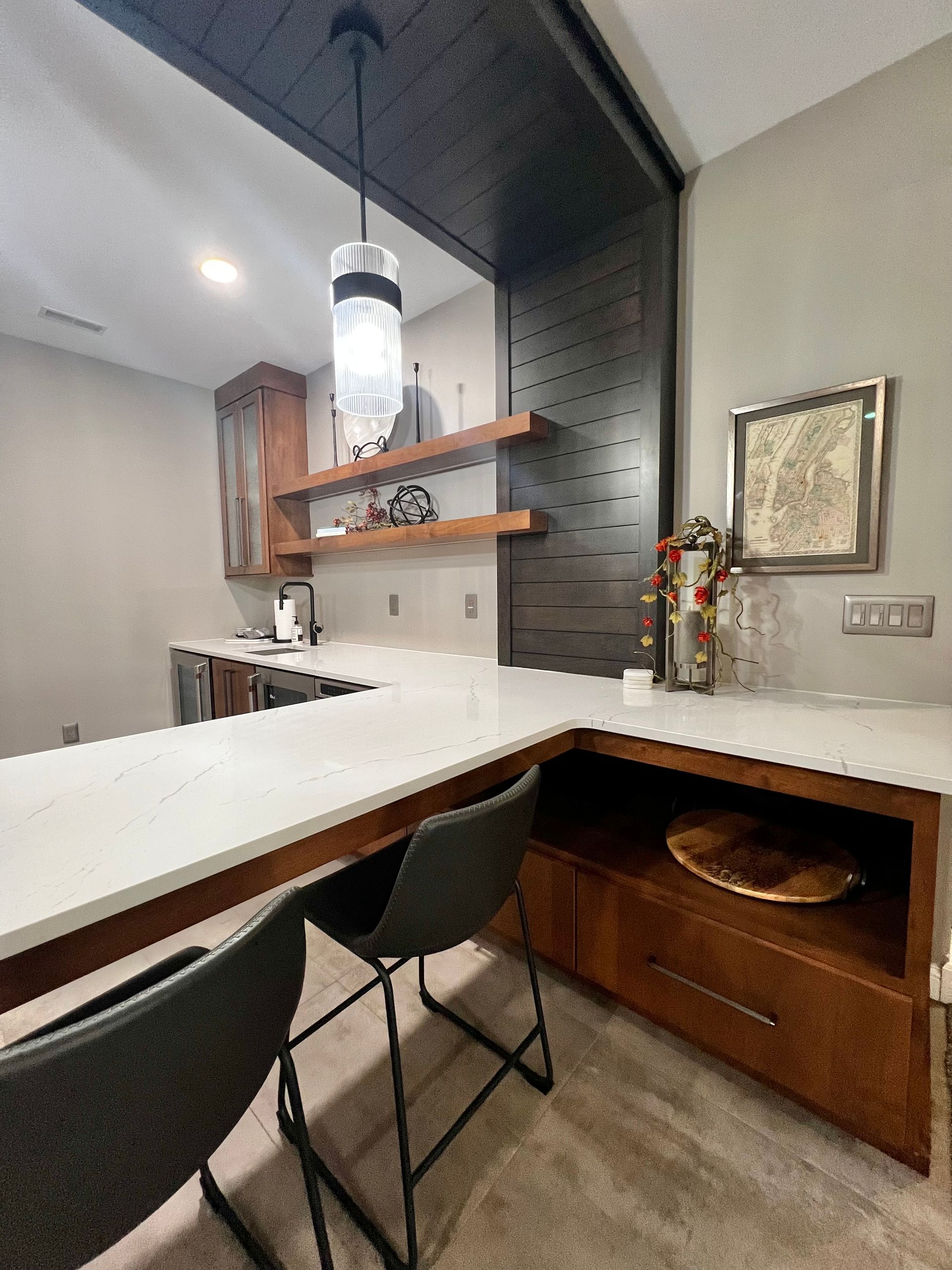 Modern home bar with white countertops, dark wood cabinetry, two black bar stools, and a decorative pendant light.