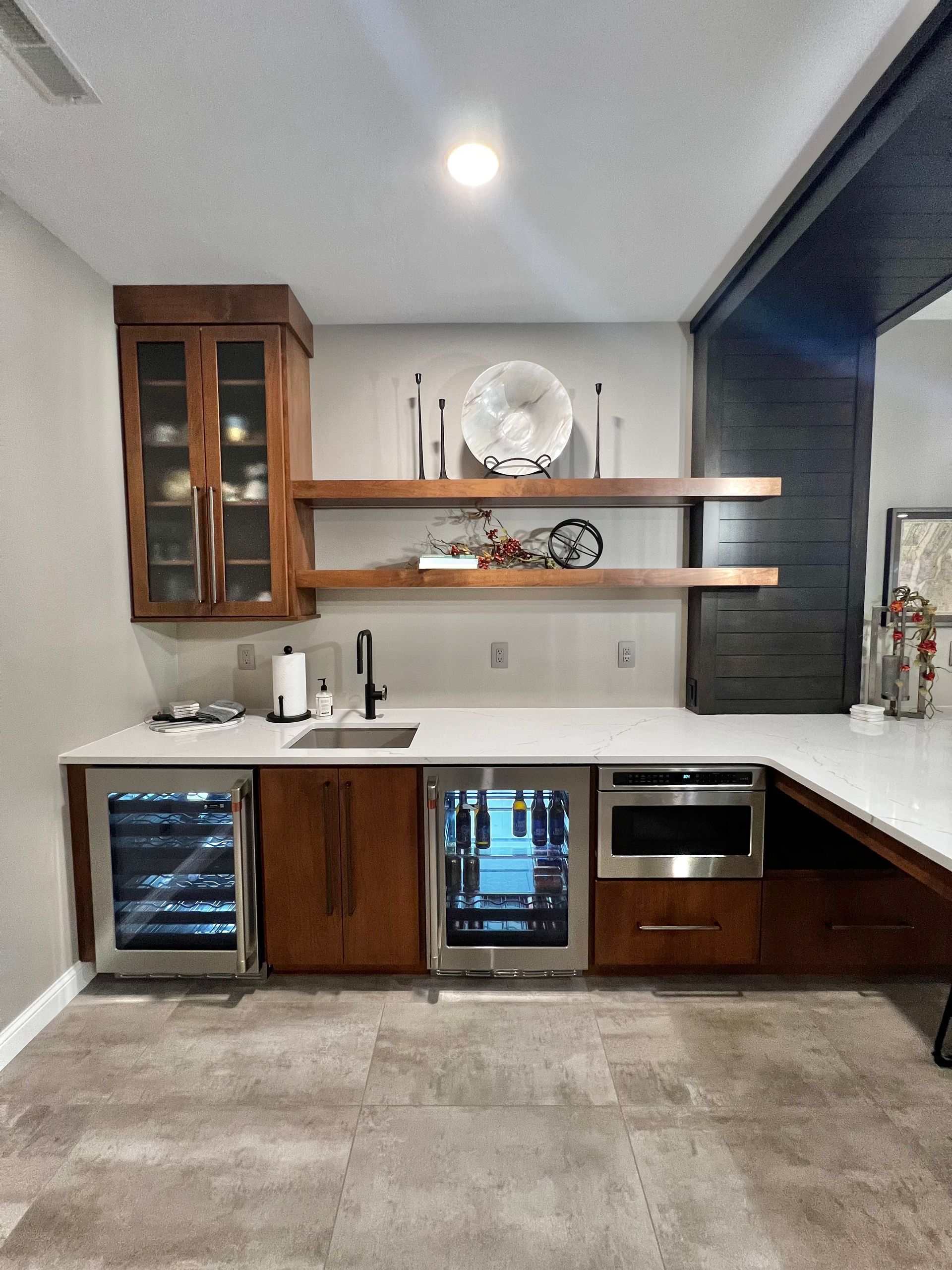 A modern wet bar featuring a wooden cabinet, open shelves, white countertops, two under-counter fridges, and a sink.
