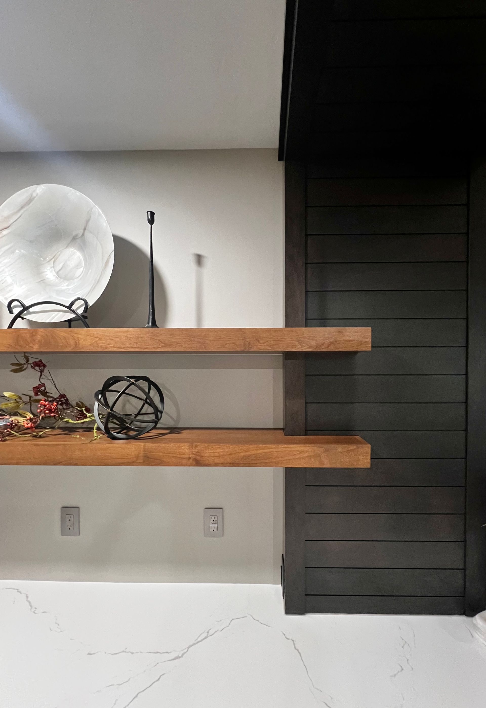 Two light wood floating shelves mounted against a gray wall and dark wood-slatted panel, with decorative items on display.