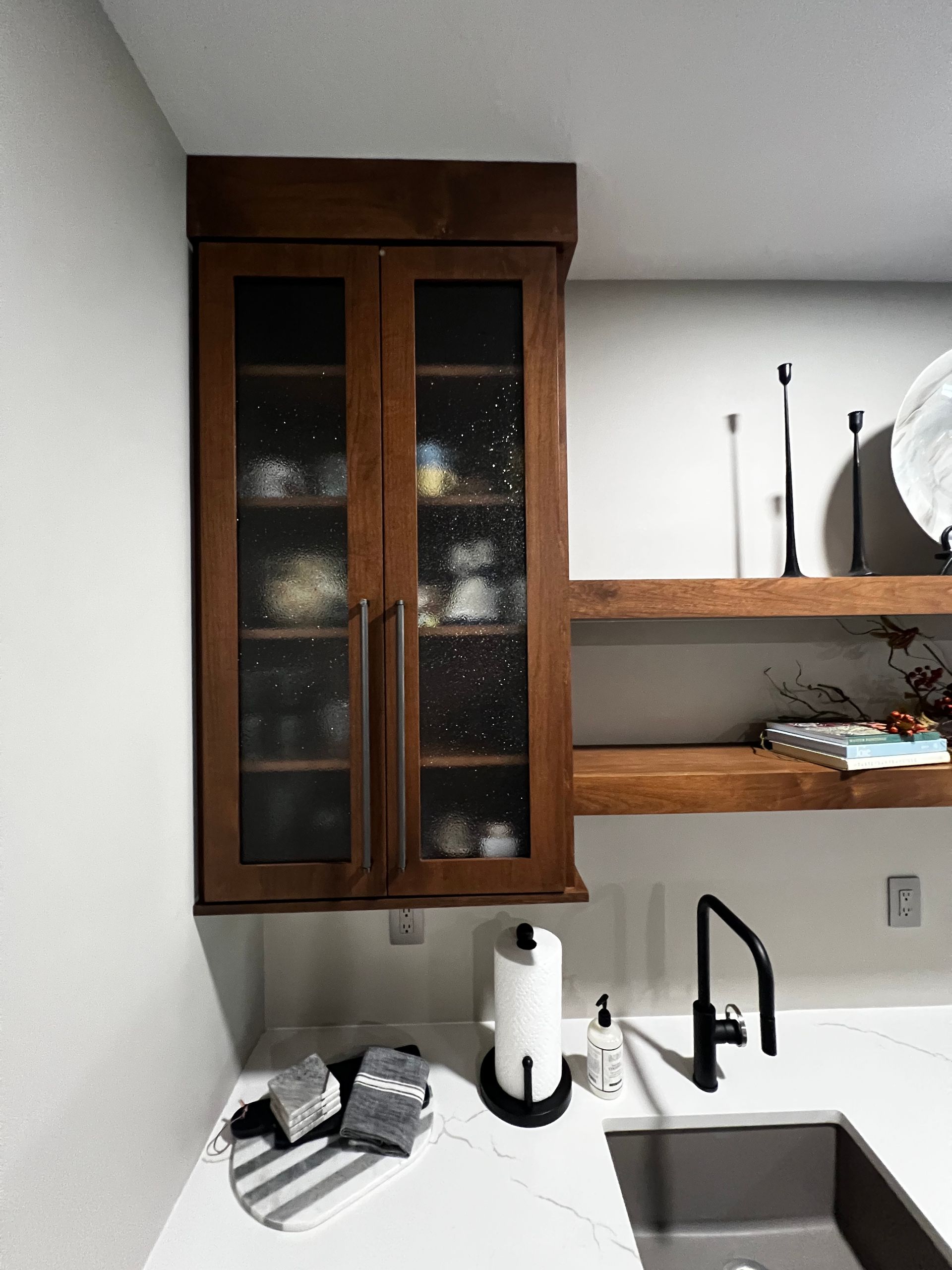 A dark wood cabinet with glass doors hangs above a white countertop, next to open shelving, a faucet, and a sink.