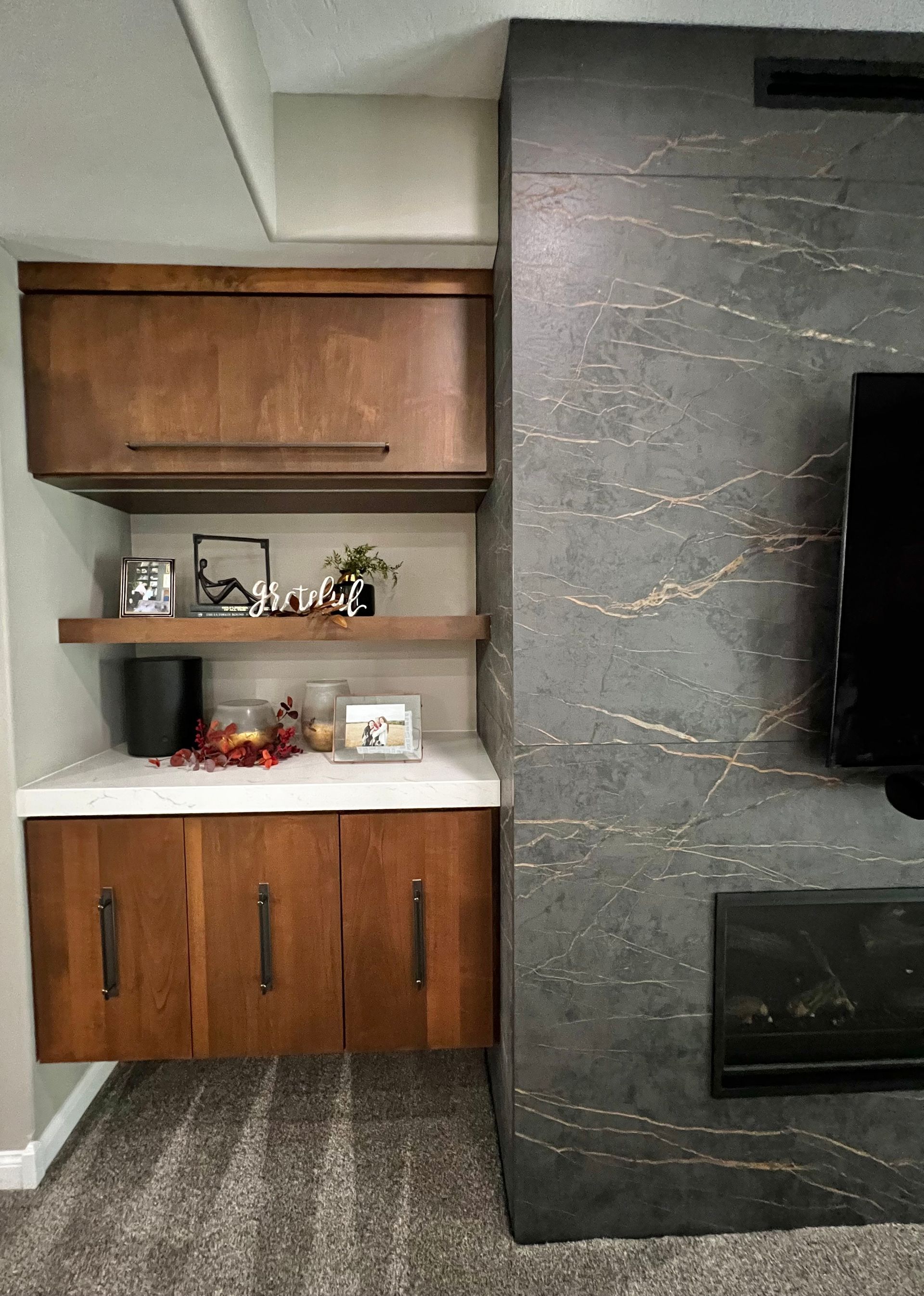 A modern floating wood cabinet with a white countertop and shelf, next to a dark gray, textured floor-to-ceiling fireplace.