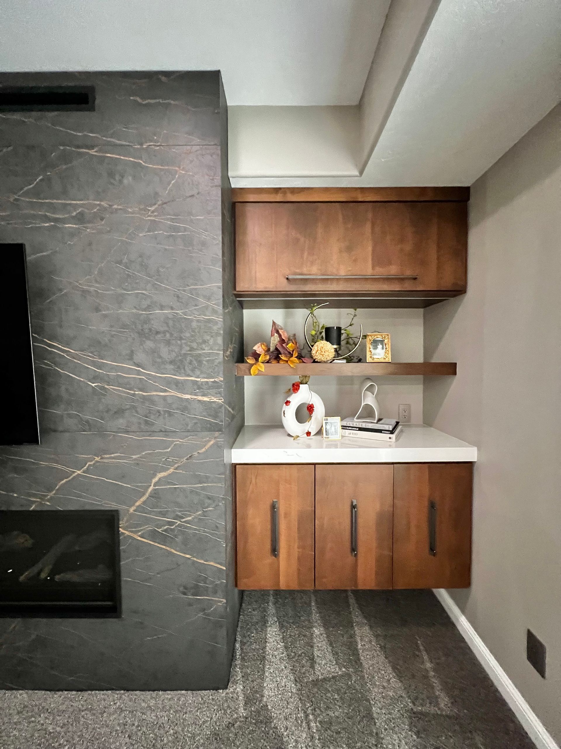 A dark gray stone fireplace sits next to a built-in wooden cabinet unit with a white countertop and display shelf.