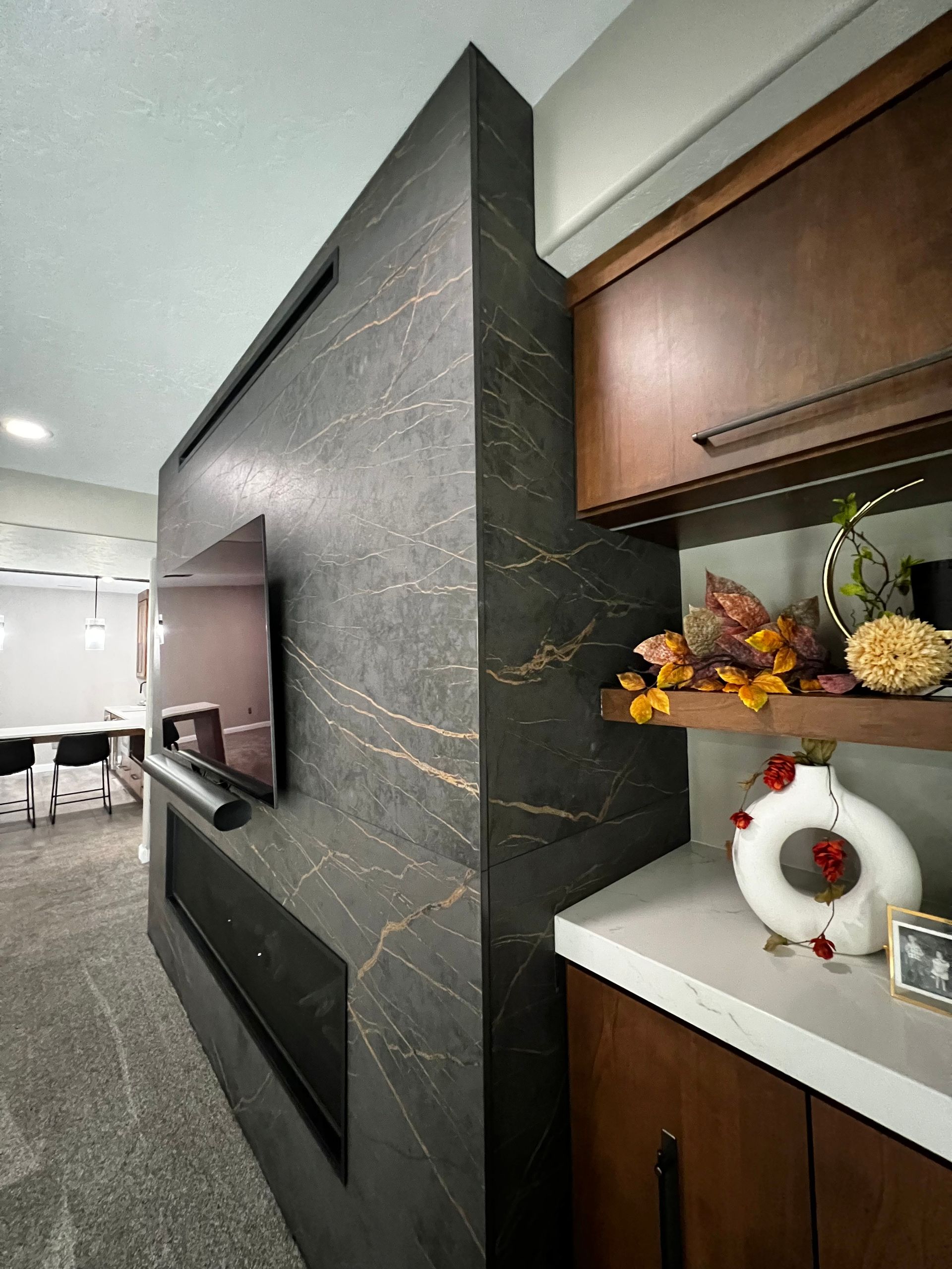A dark, veined stone fireplace with a wall-mounted TV, adjacent to wood cabinets and a shelf with decorative items.