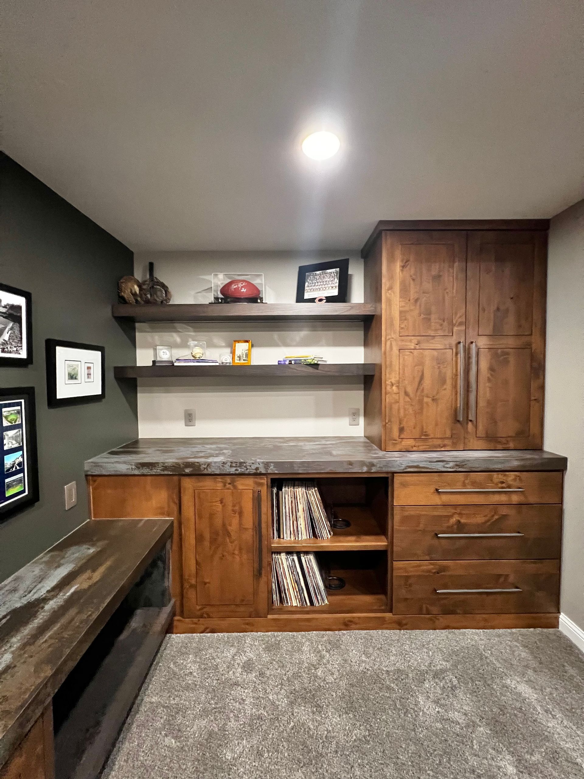 A basement home bar area with wood cabinets, concrete counters, floating shelves, and a gray-painted accent wall.