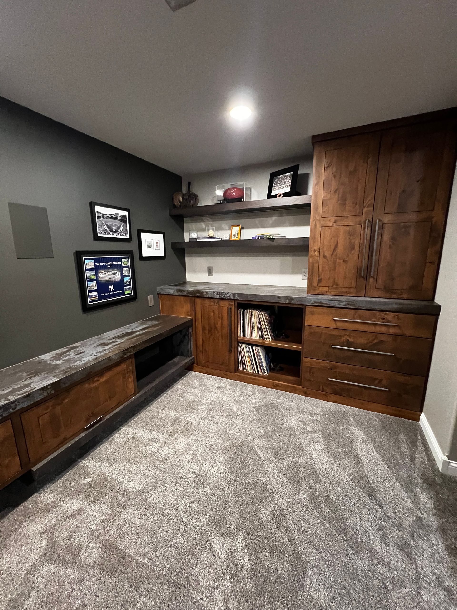 A basement room with dark gray walls, custom dark wood cabinetry, a long bench, open shelves, and gray carpeted floors.