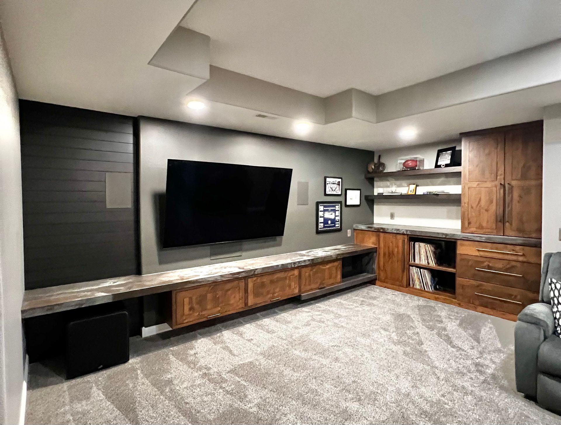 A basement media room featuring a large mounted television, floating wooden cabinetry, open shelving, and gray carpeting.