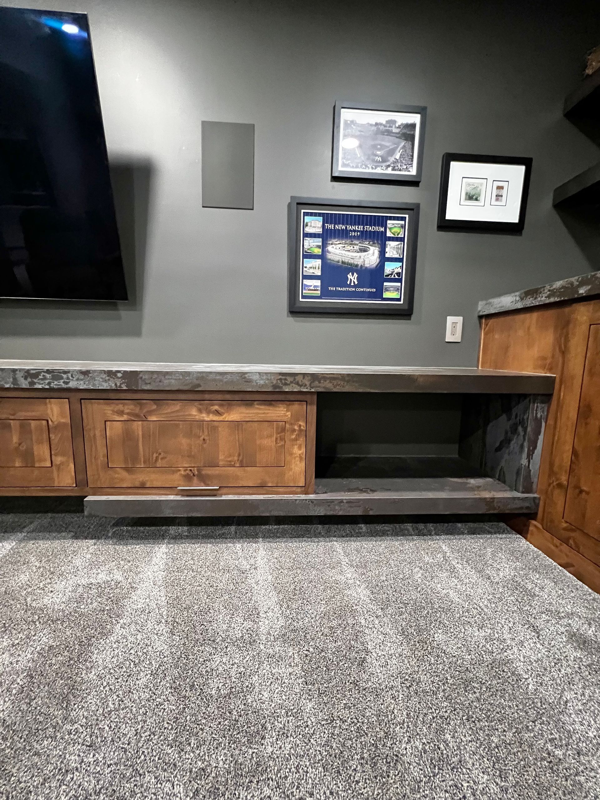 A low wooden entertainment console with an open storage cubby sits against a dark gray wall beneath a TV and framed art.