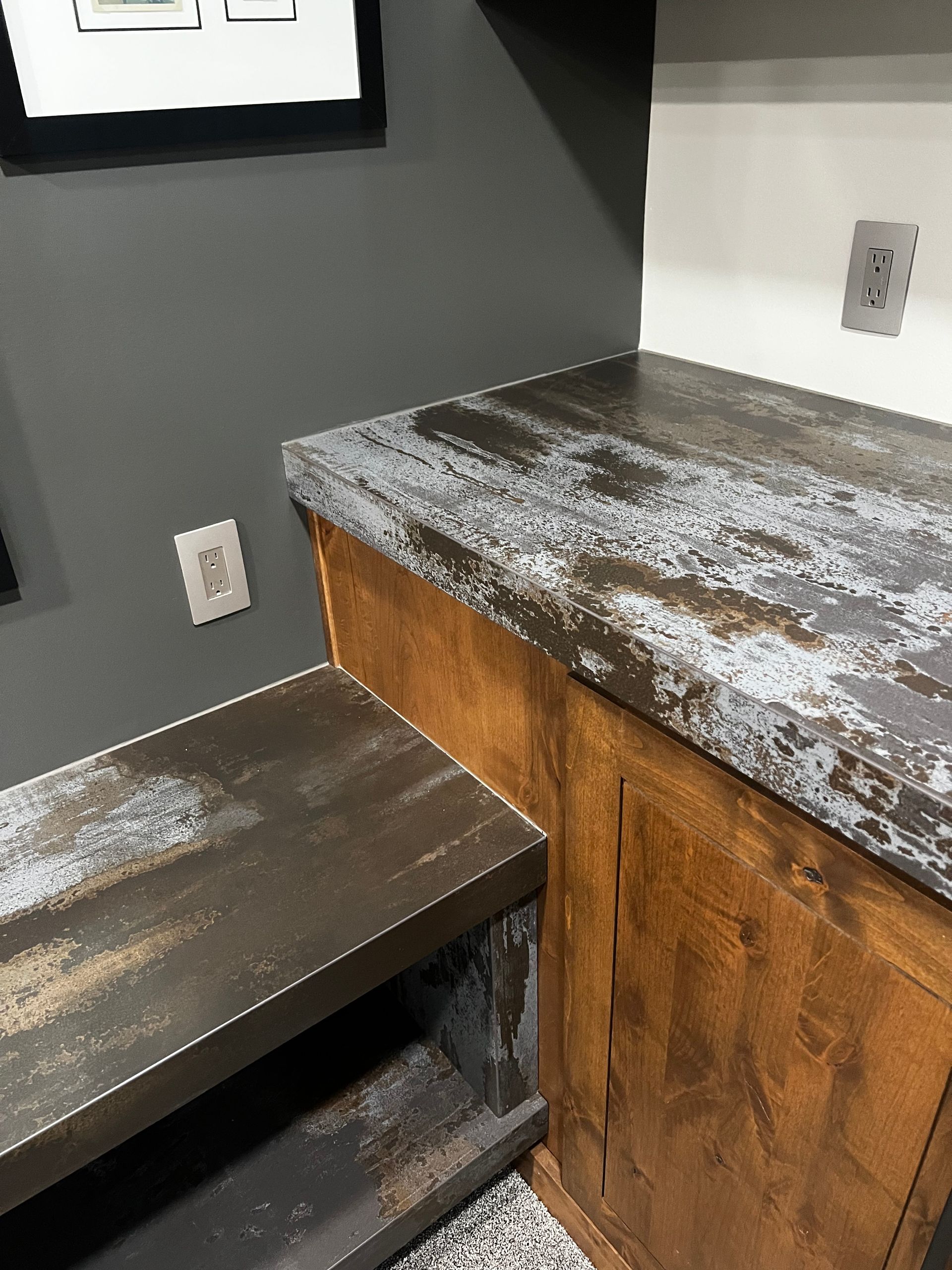 A rustic wooden cabinet with a distressed gray countertop and lower shelf sits against a dark gray wall with outlets.