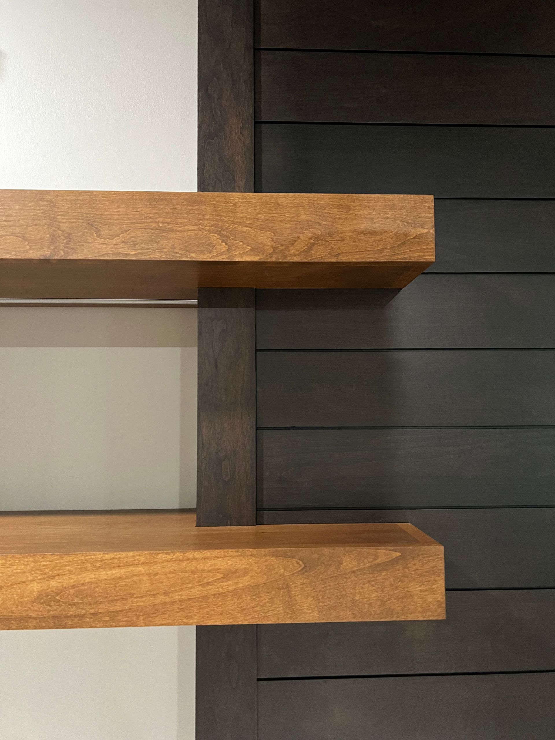 Two wooden floating shelves mounted on a vertical dark wood post against a gray wall and dark shiplap paneling.
