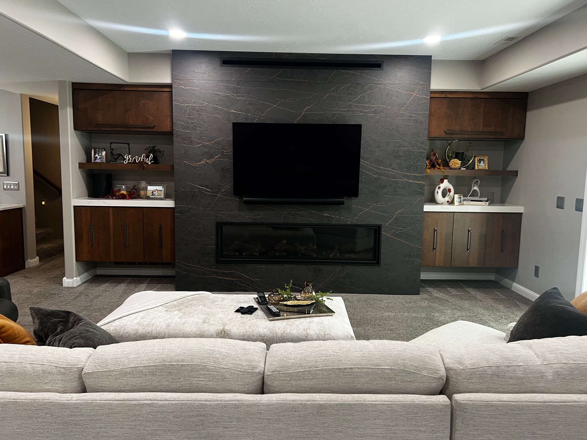 A modern living room with a large, dark stone fireplace wall, wall-mounted TV, wood cabinetry, and a light-colored sofa.