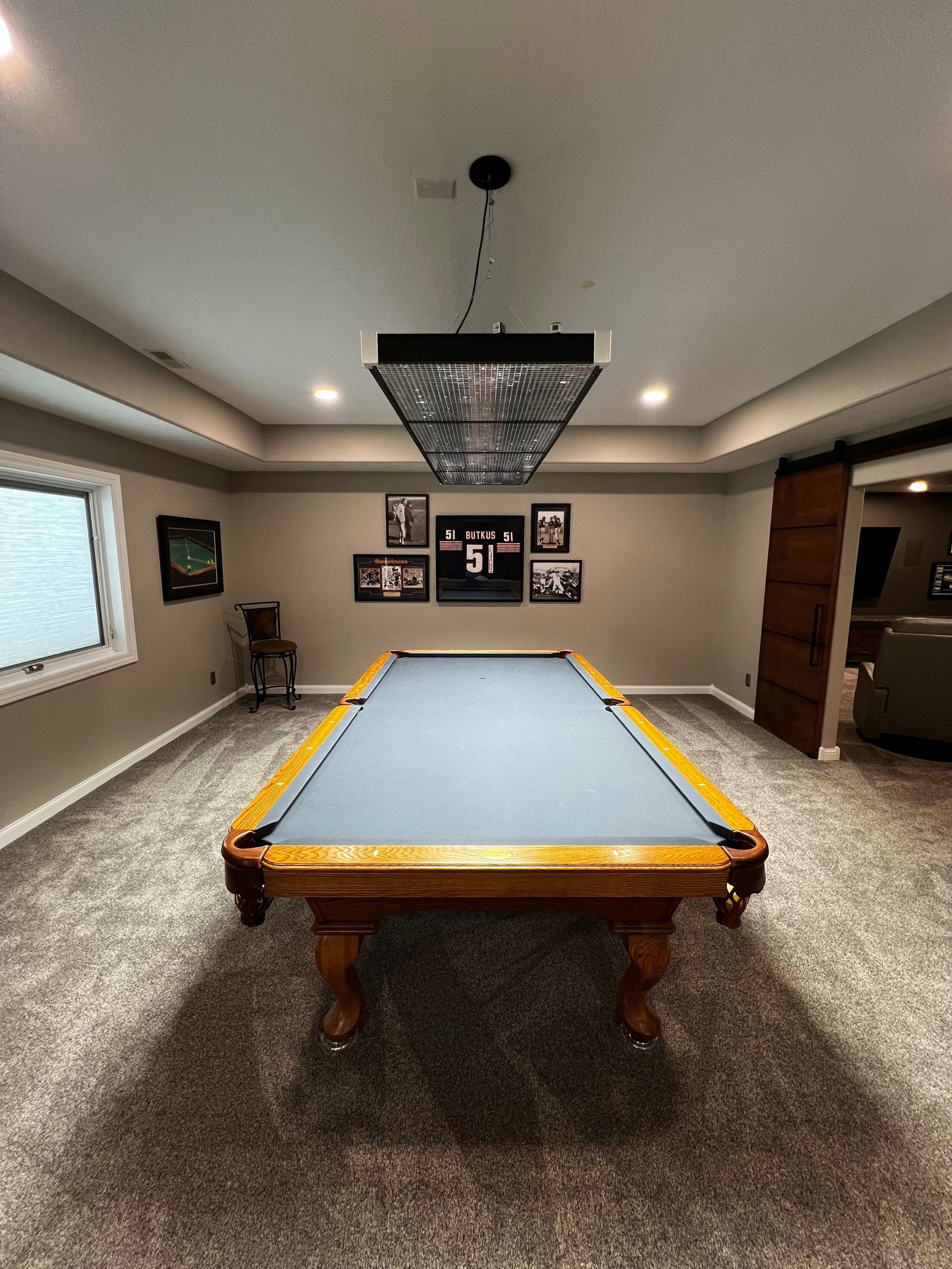 A basement game room featuring a billiards table with blue felt, a modern rectangular light fixture, and framed wall art.