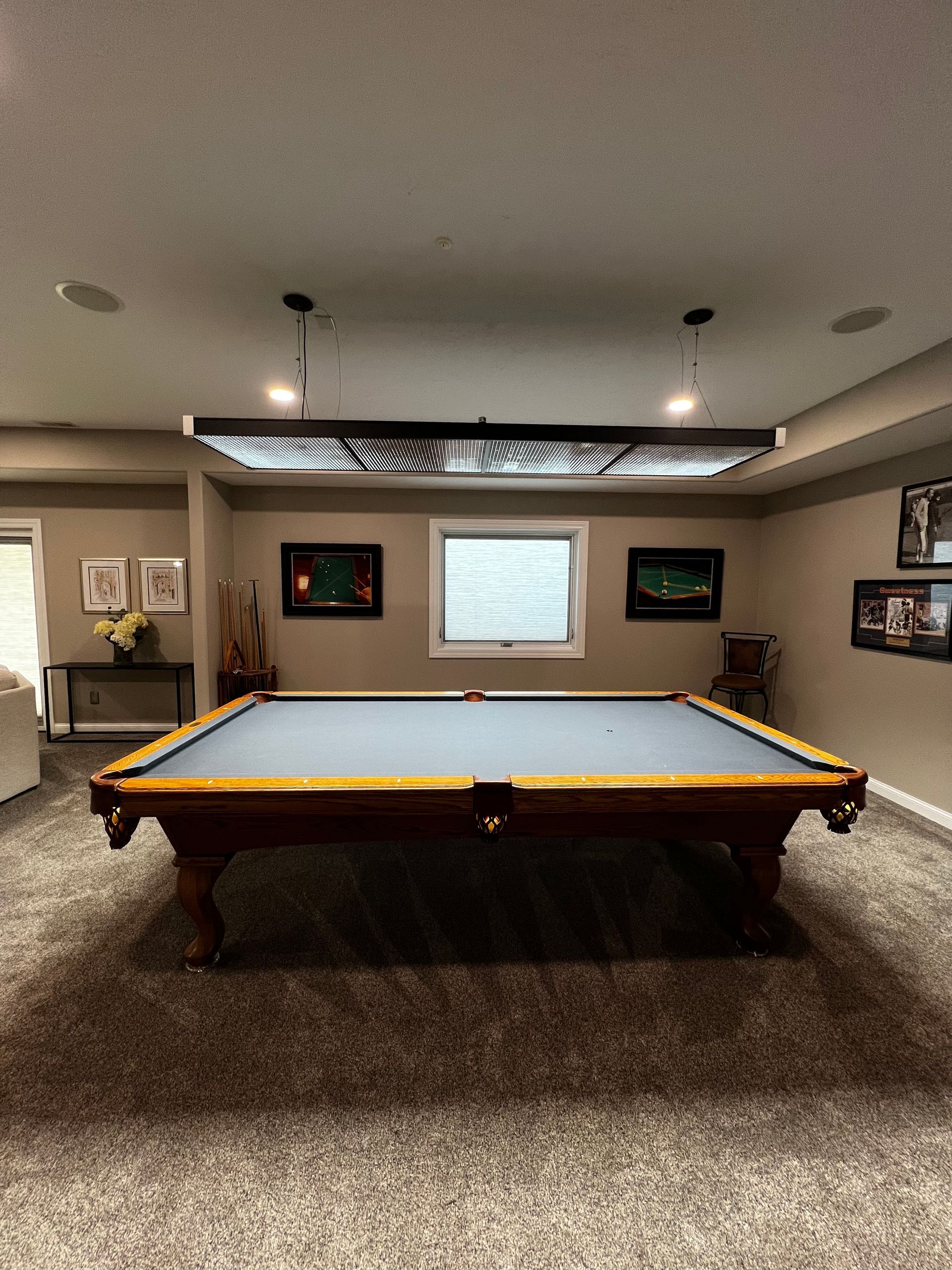 A billiards table with a grey felt surface in a carpeted basement room, illuminated by a large modern light fixture.