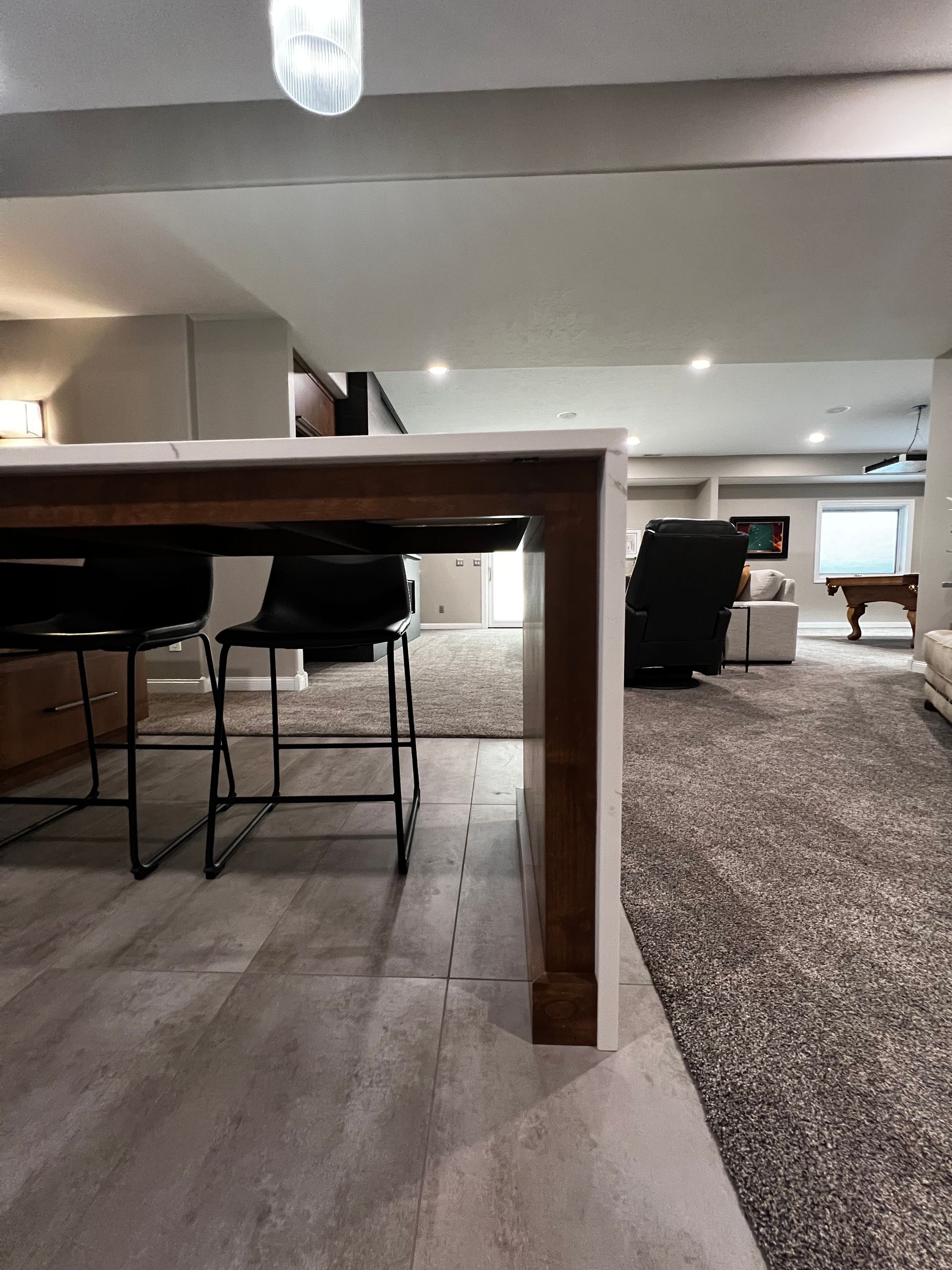 A view from under a wood and white kitchen island, looking out onto a carpeted basement with stools, seating, and a pool table.