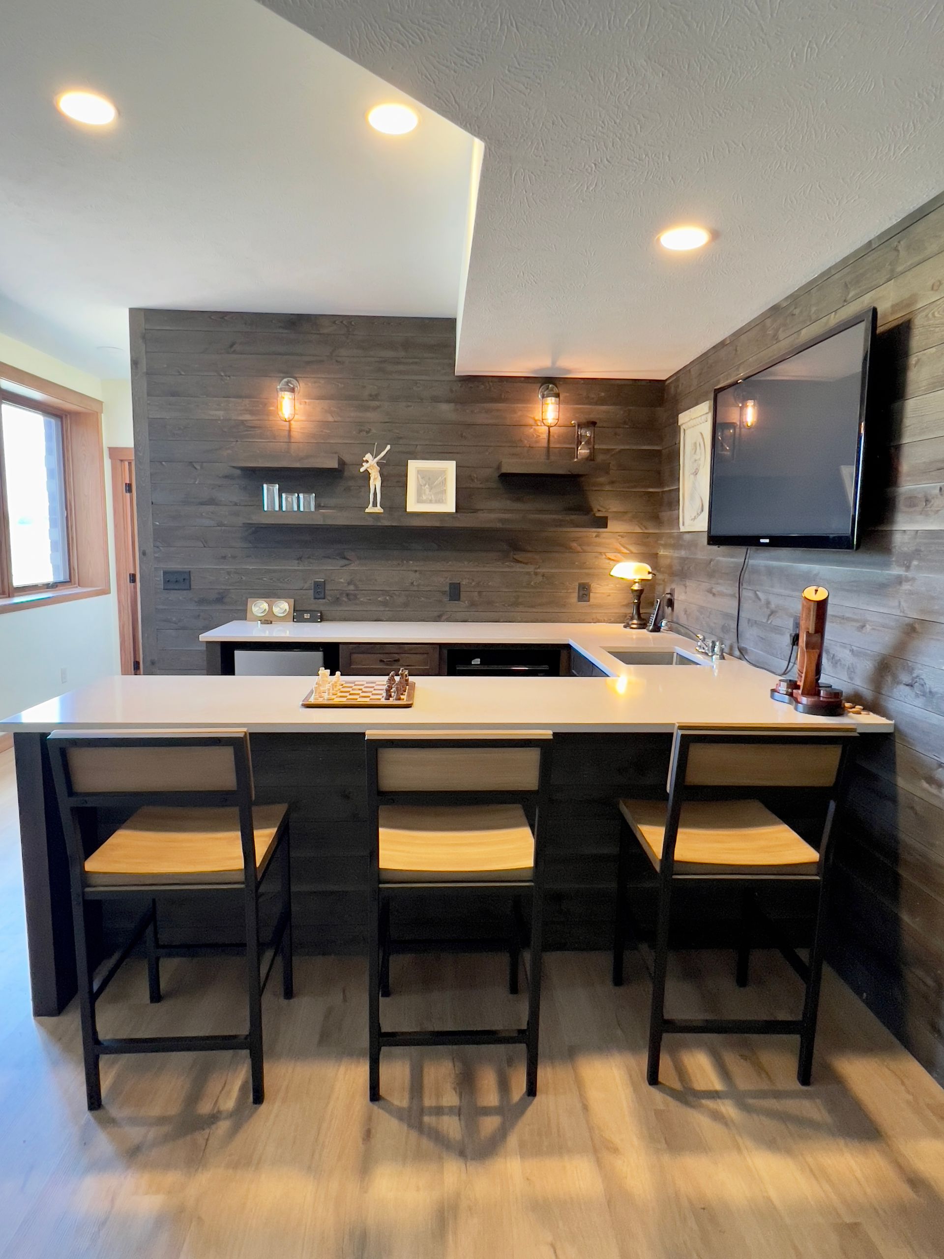 A modern home bar with a white counter, three bar stools, dark wood paneling, open shelving, and a wall-mounted TV.