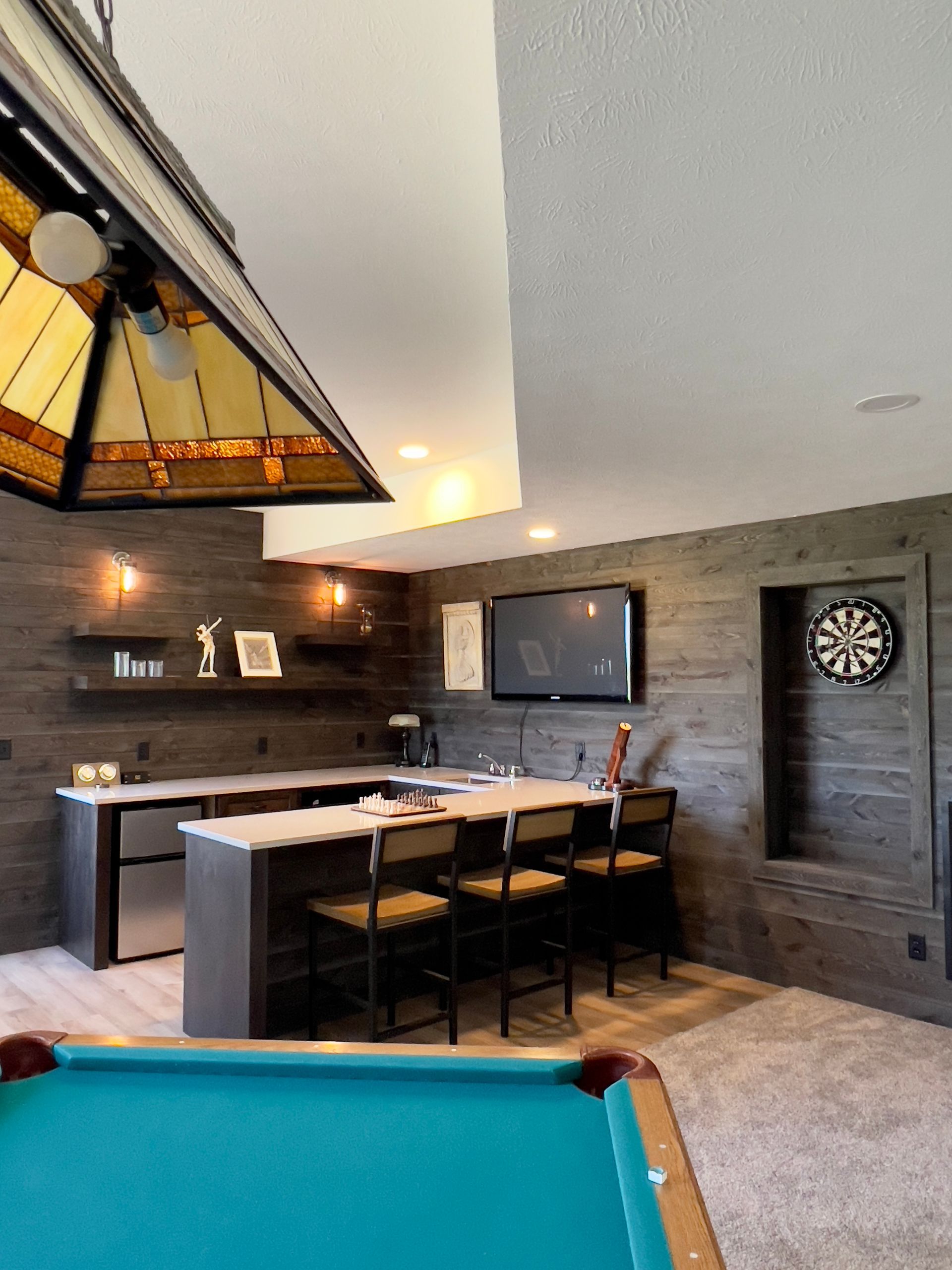 A finished basement featuring a pool table, bar seating, dark wood-paneled walls, a wall-mounted TV, and a dartboard.