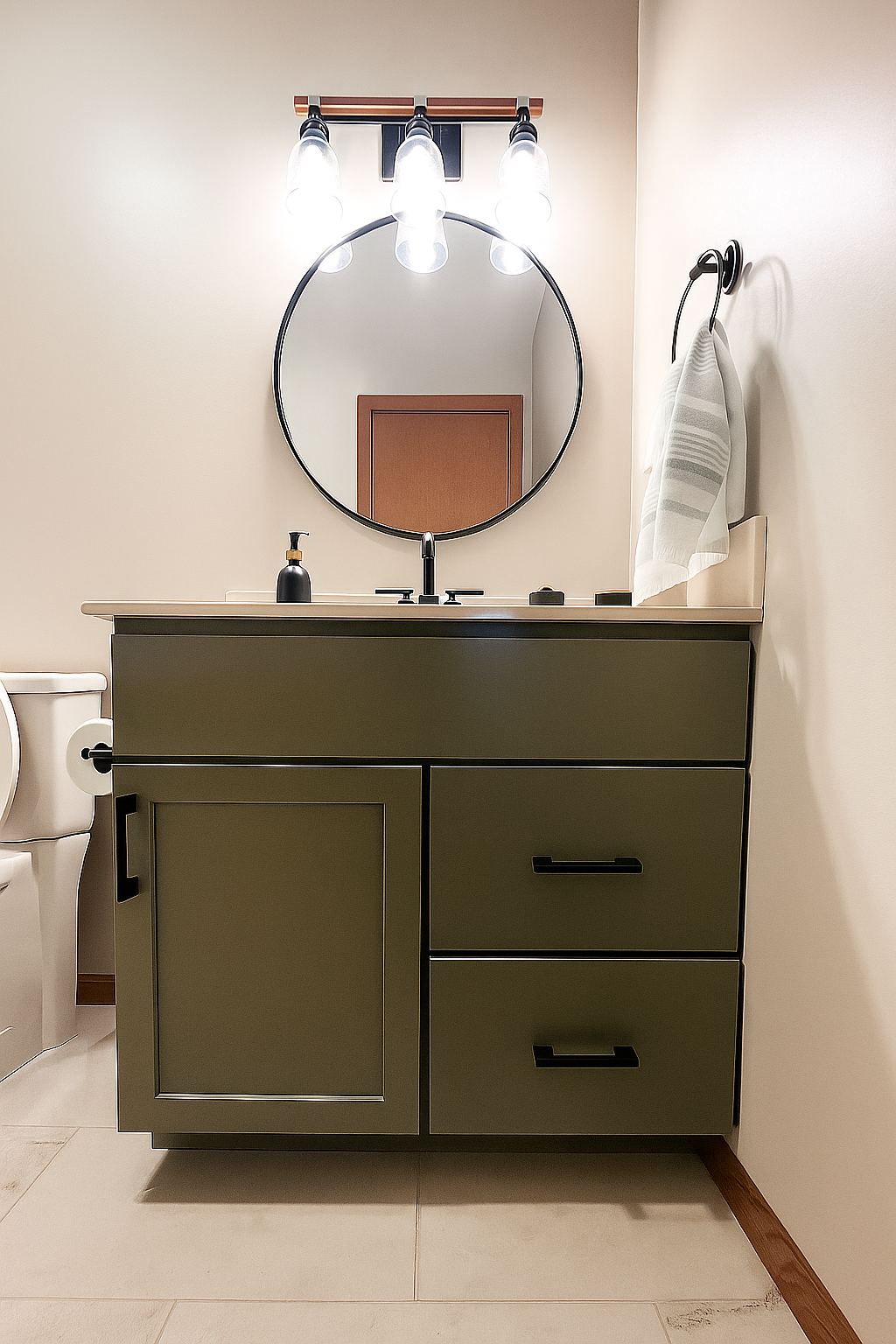 A bathroom vanity with an olive-green cabinet, matte black hardware, a round mirror, and a three-light vanity fixture.