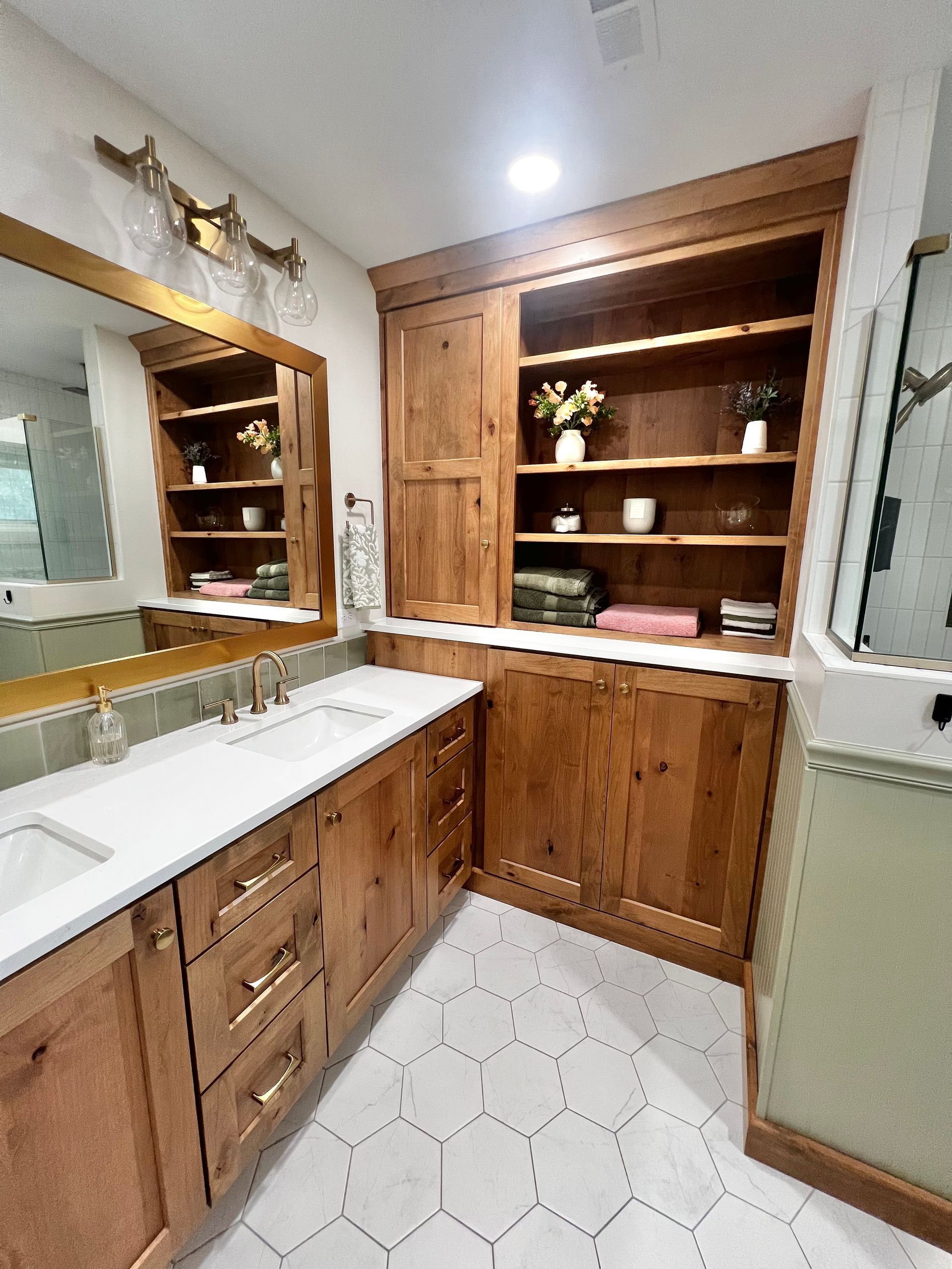 A rustic-style bathroom with wooden cabinetry, open shelving, white hexagonal tile floors, and a large vanity mirror.