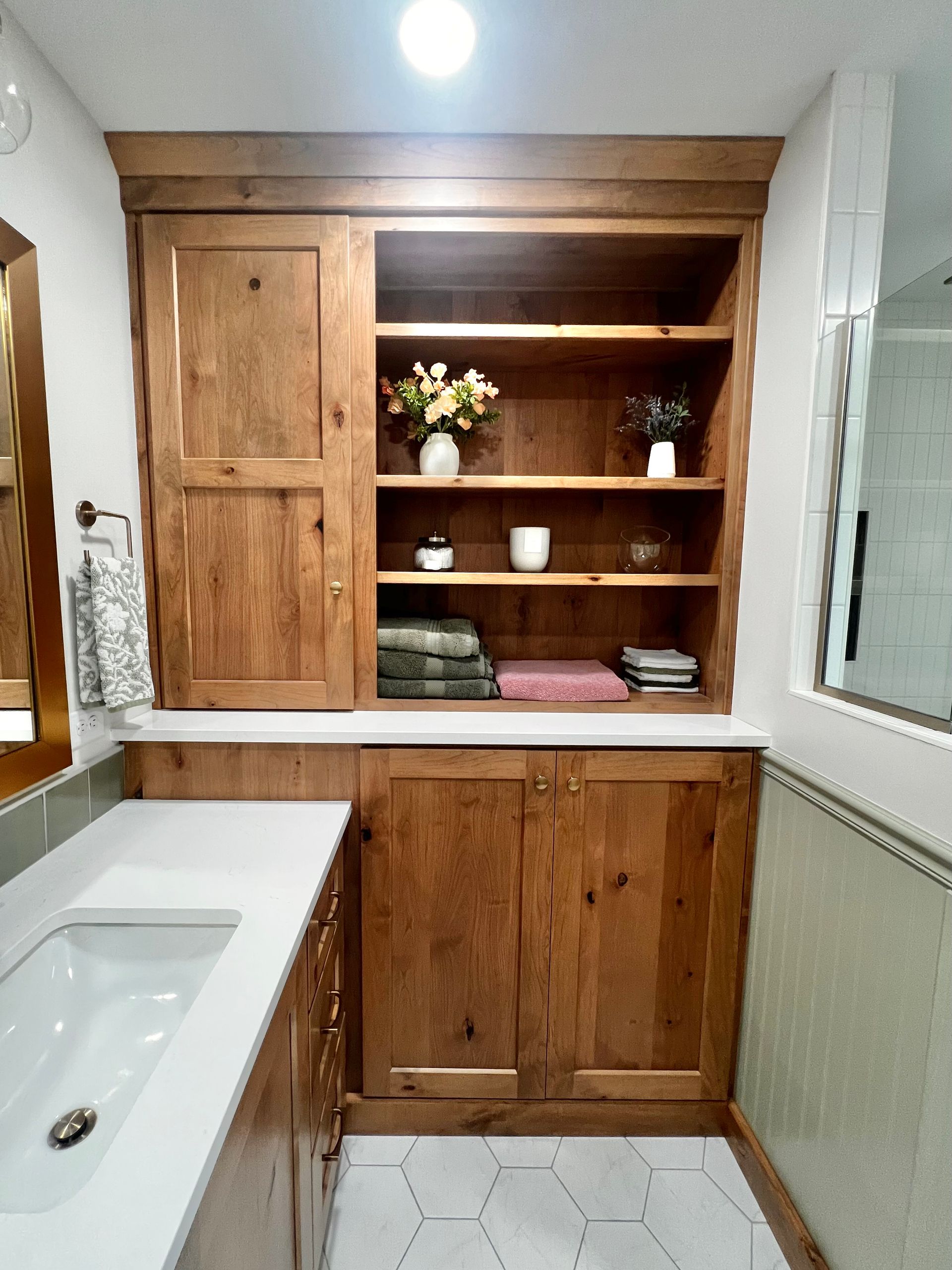 A wooden bathroom vanity with open shelving, a white countertop, an integrated sink, and white hexagonal floor tiles.