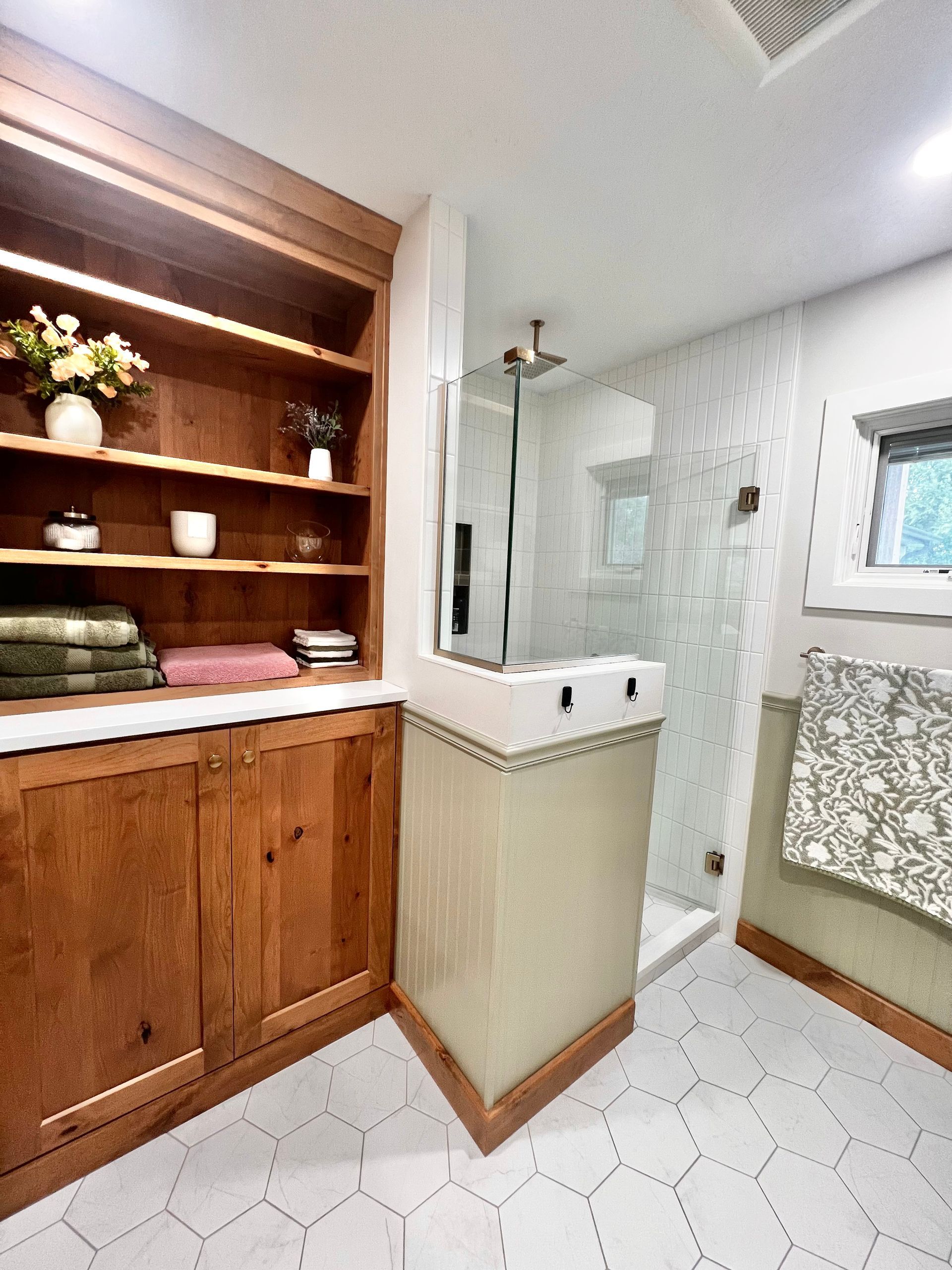 A bathroom featuring a wooden storage unit, a glass shower stall with light green tile walls, and white hexagon floor tiles.