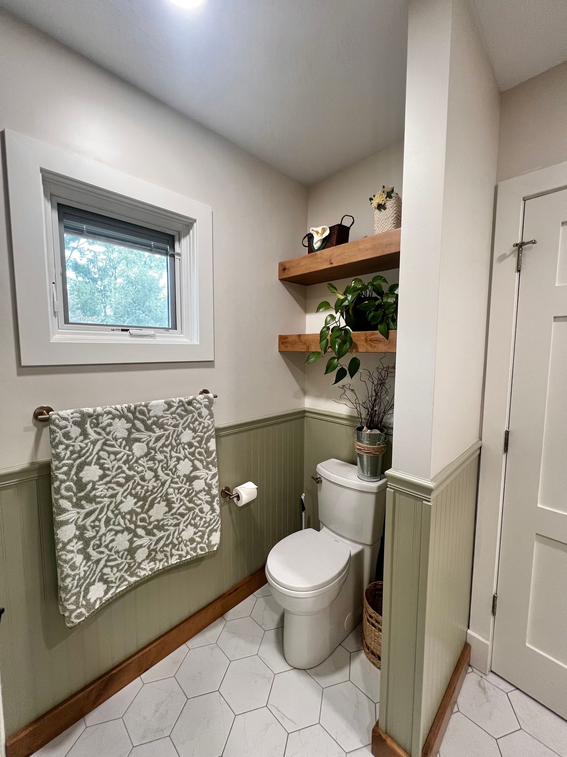 A small bathroom with sage-green wainscoting, white hexagonal floor tiles, a window, and two wooden shelves above a toilet.