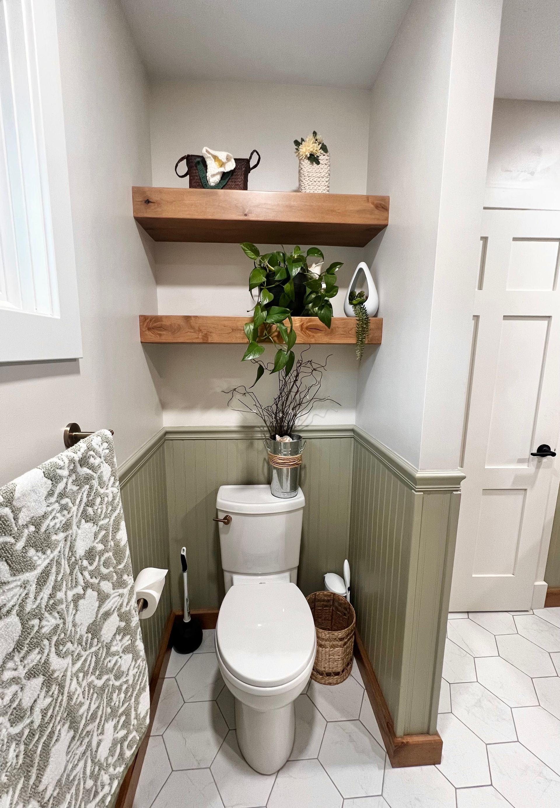 A small bathroom with sage green wainscoting, white hex floor tiles, and two wood shelves above a white toilet.
