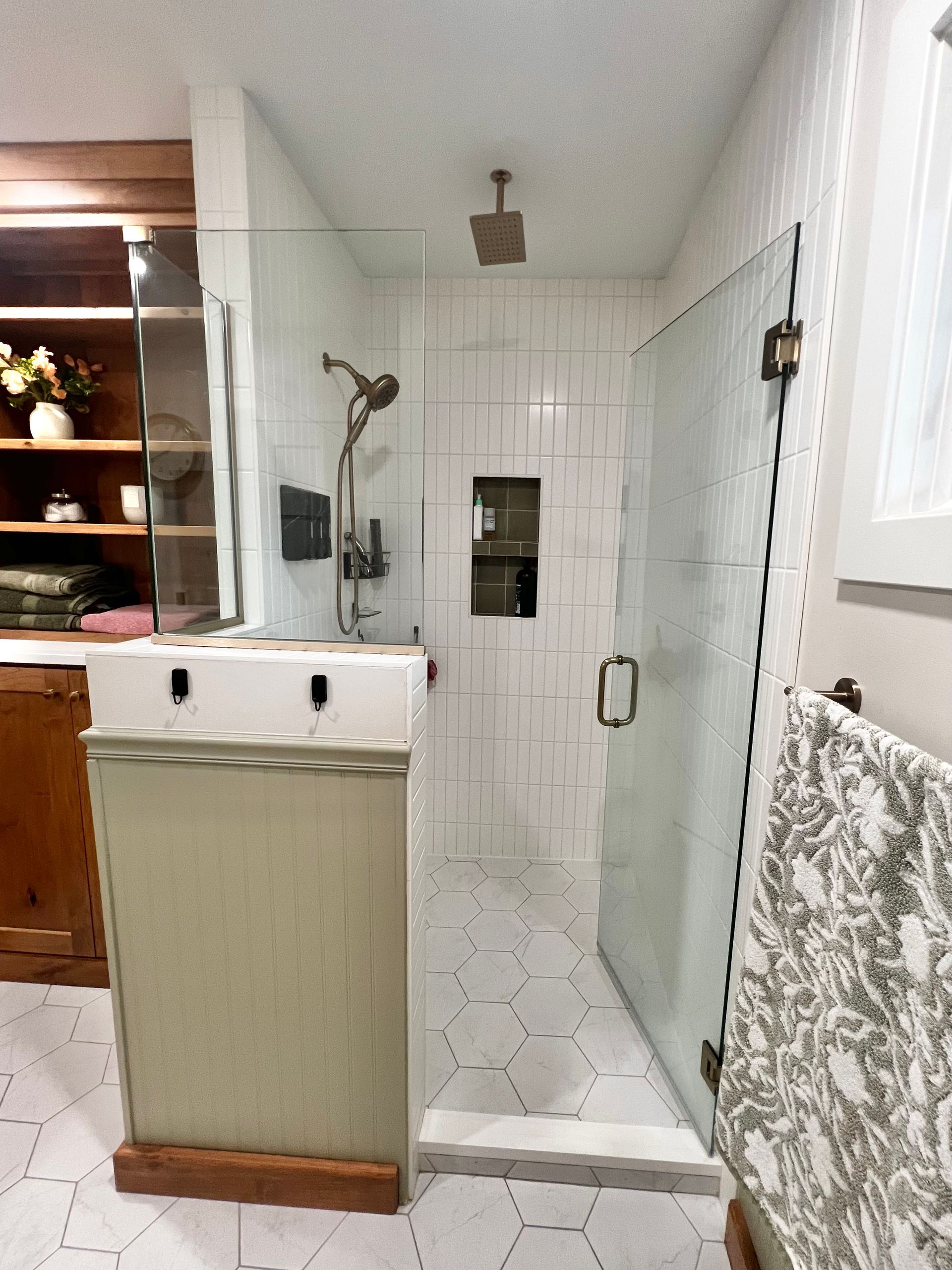 A modern walk-in shower with white tiled walls, a glass door, rainfall showerhead, and a separate handheld sprayer.