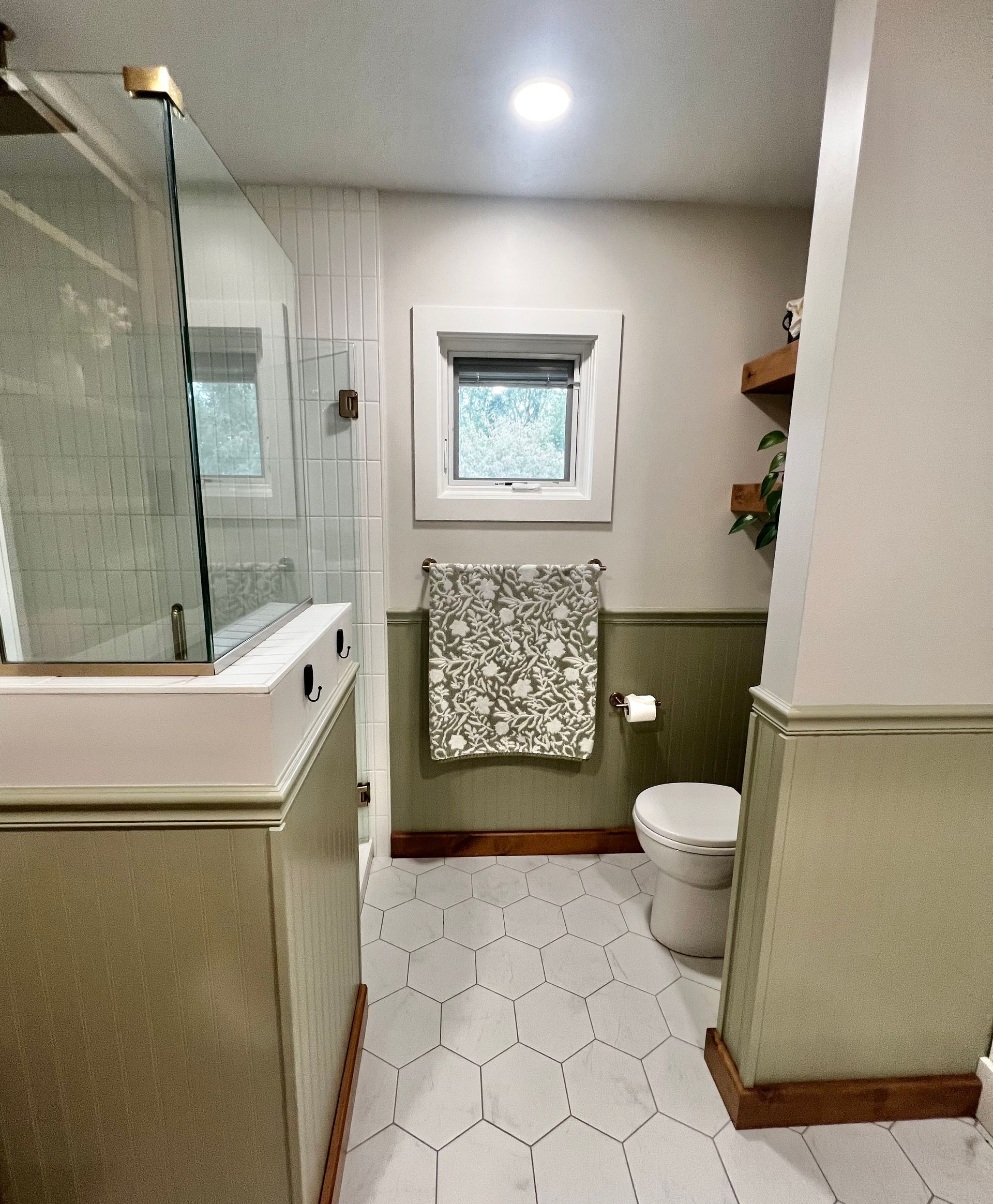 A modern bathroom with sage green wainscoting, white hexagonal floor tiles, a glass shower, and a window with a towel.