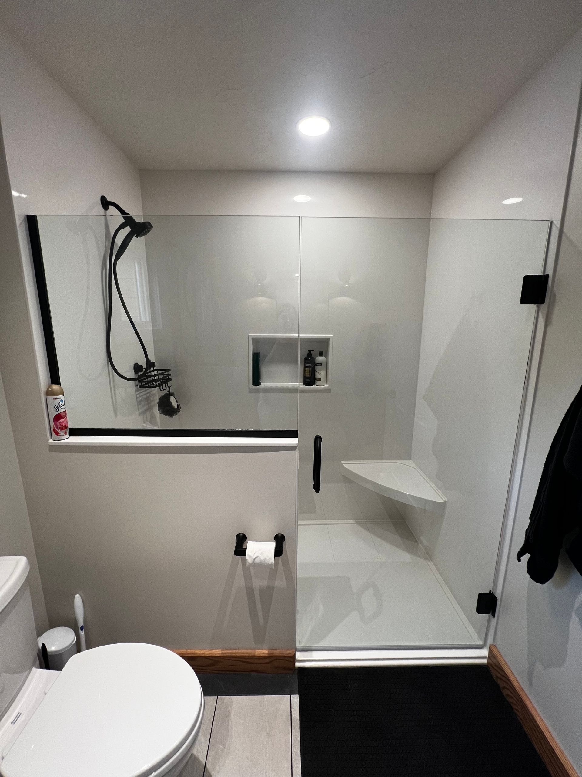 A bathroom features a walk-in shower with a glass enclosure, a built-in corner seat, wall shelving, and black fixtures.