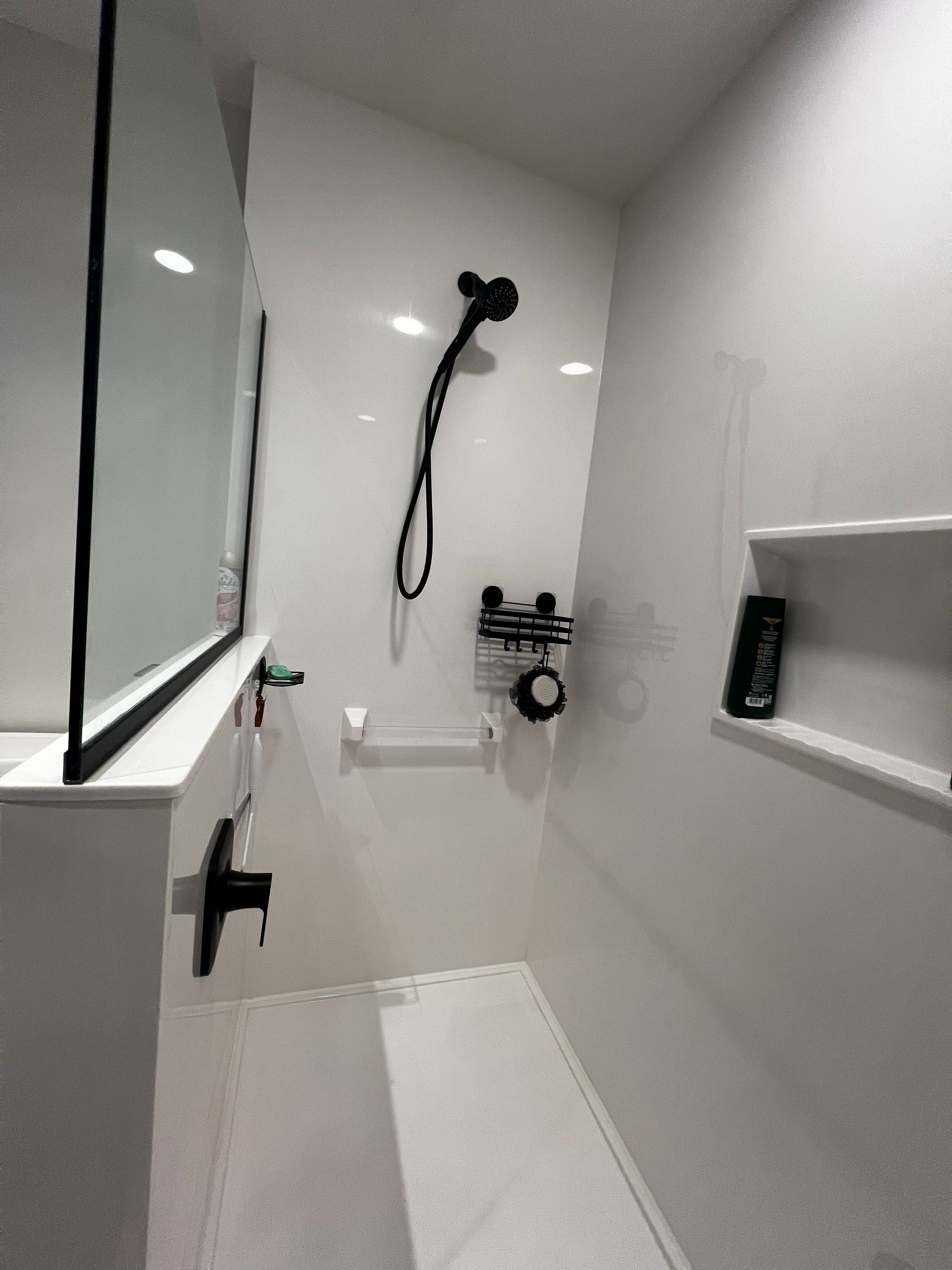 A white walk-in shower with a black handheld showerhead, wall-mounted faucet, grab bar, and a recessed shelving niche.