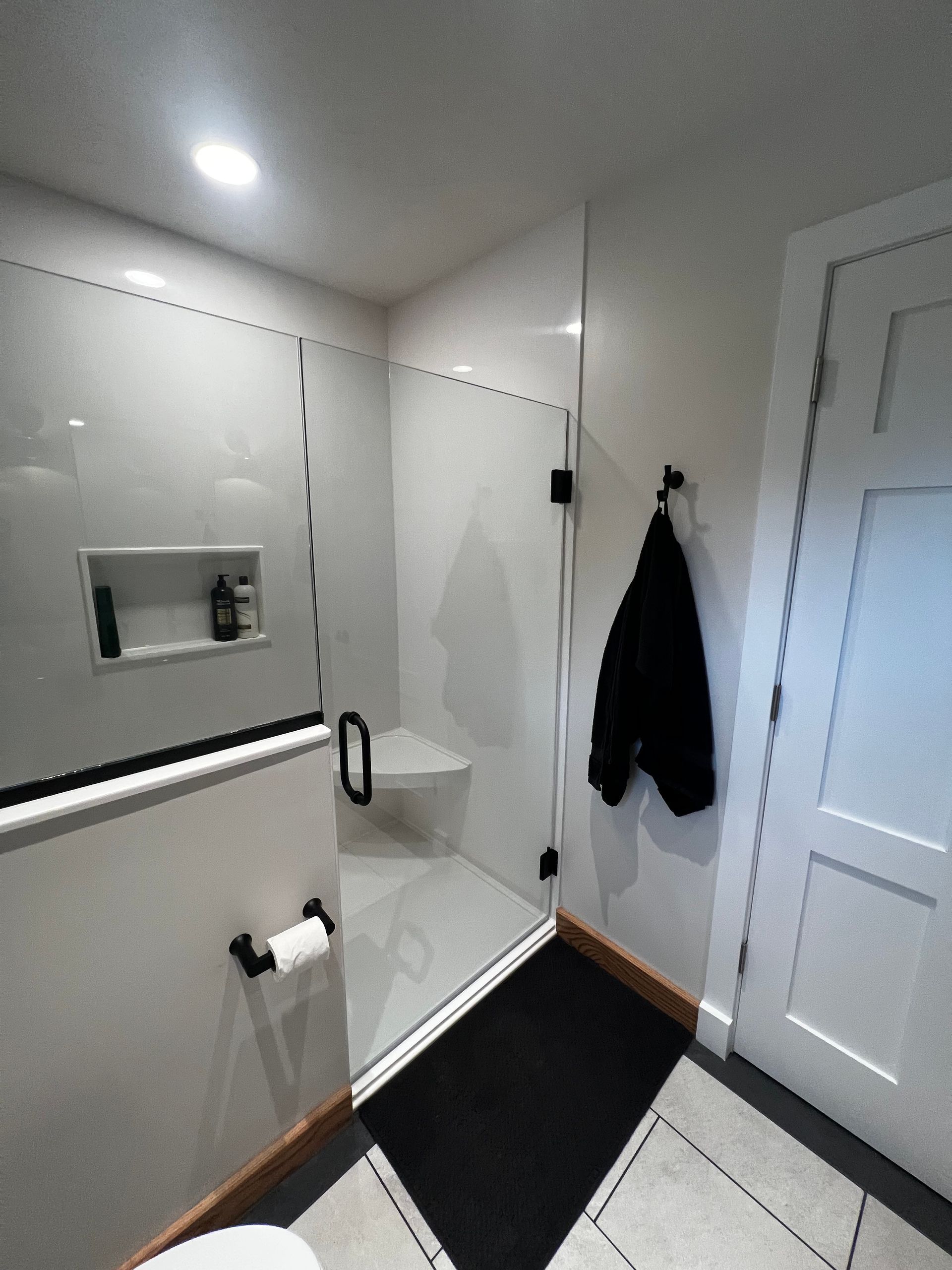A walk-in shower with white tiled walls, a corner seat, and a glass door, adjacent to a white door and a toilet paper holder.