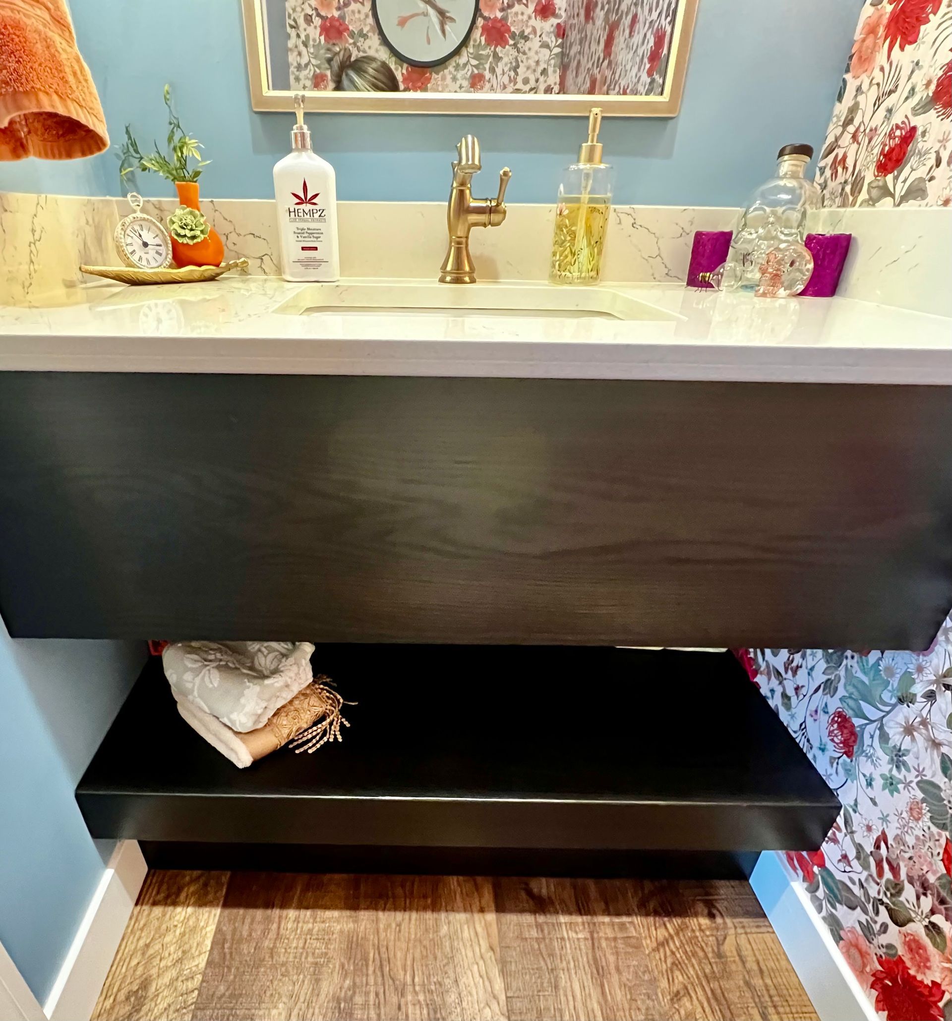 A bathroom vanity with a dark wood cabinet, white countertop, gold faucet, and a lower shelf holding folded white towels.