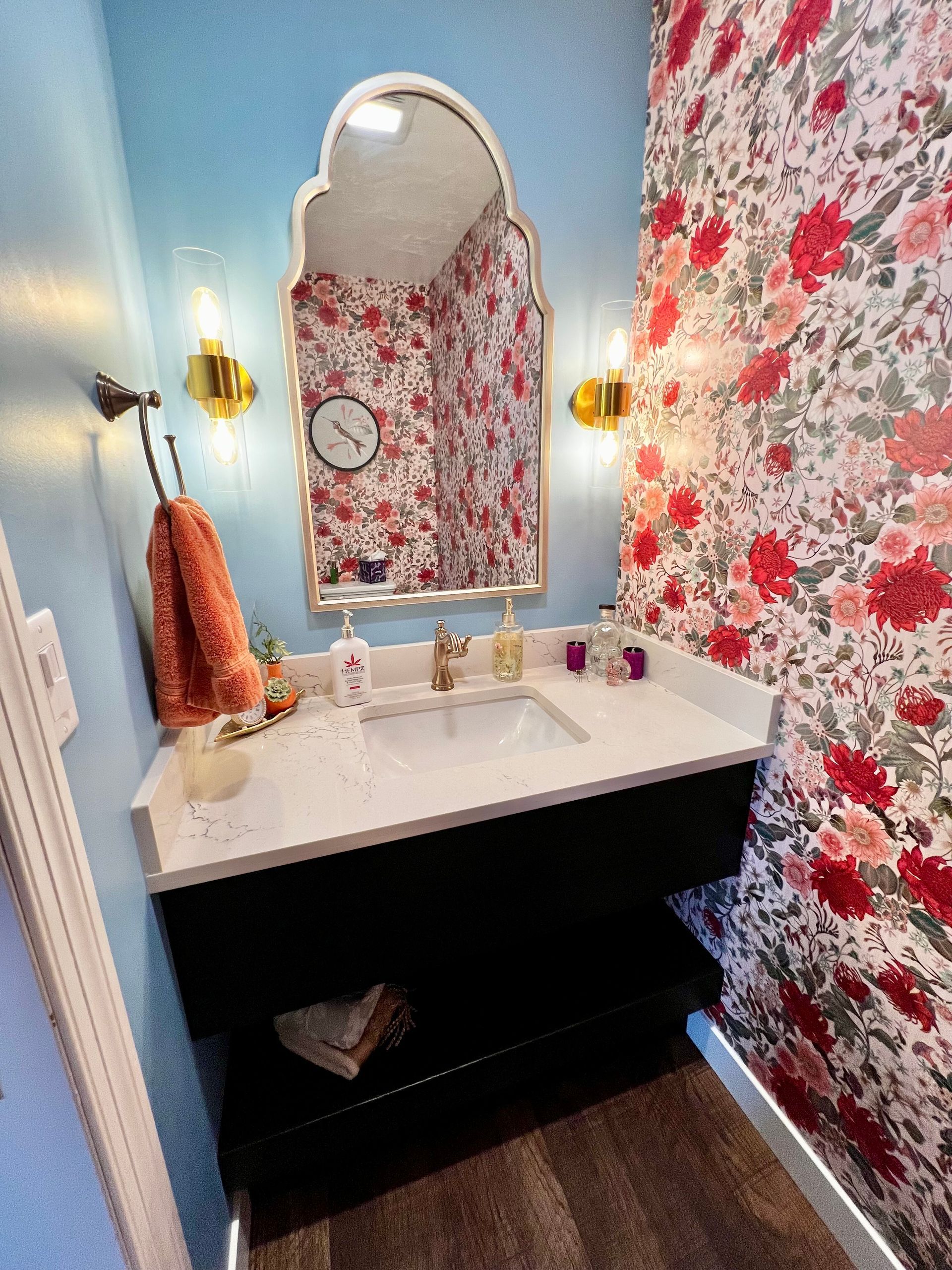 A bathroom vanity with a black cabinet, white countertop, sink, and an arched mirror, set against floral wallpaper.