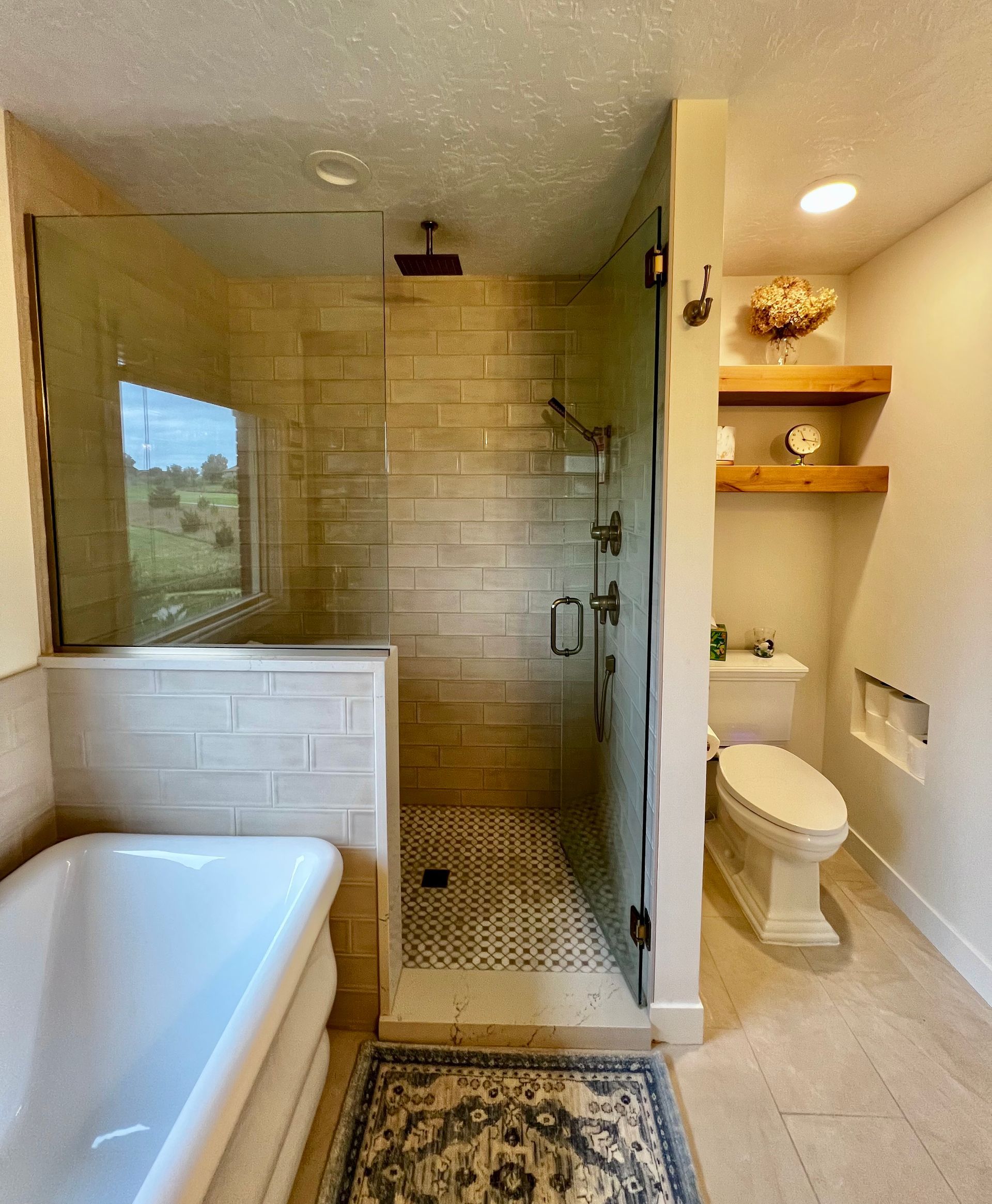A bathroom with a freestanding white tub, a glass-enclosed shower with tiled walls, and a toilet with wooden shelves.