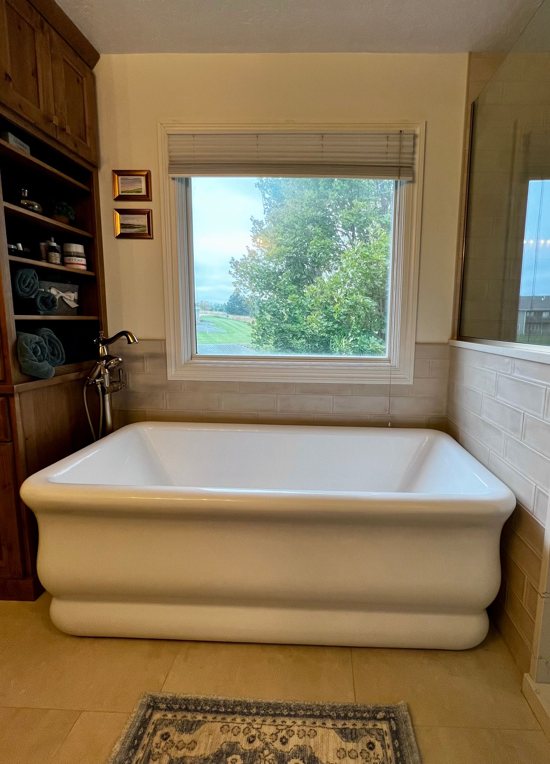 A white, rectangular soaking bathtub sits in front of a window with a view of trees, next to wooden shelving.