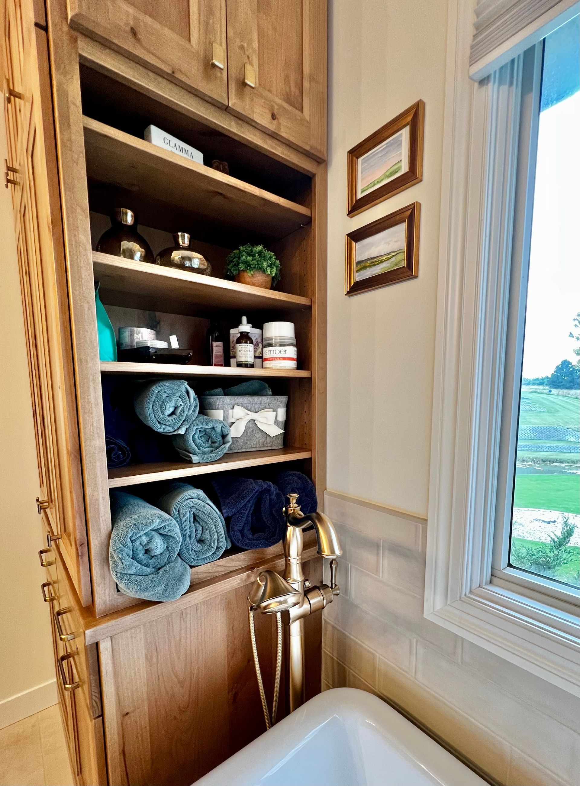 Tall wooden shelving unit in a bathroom filled with rolled blue towels, toiletries, and decor next to a window and tub.