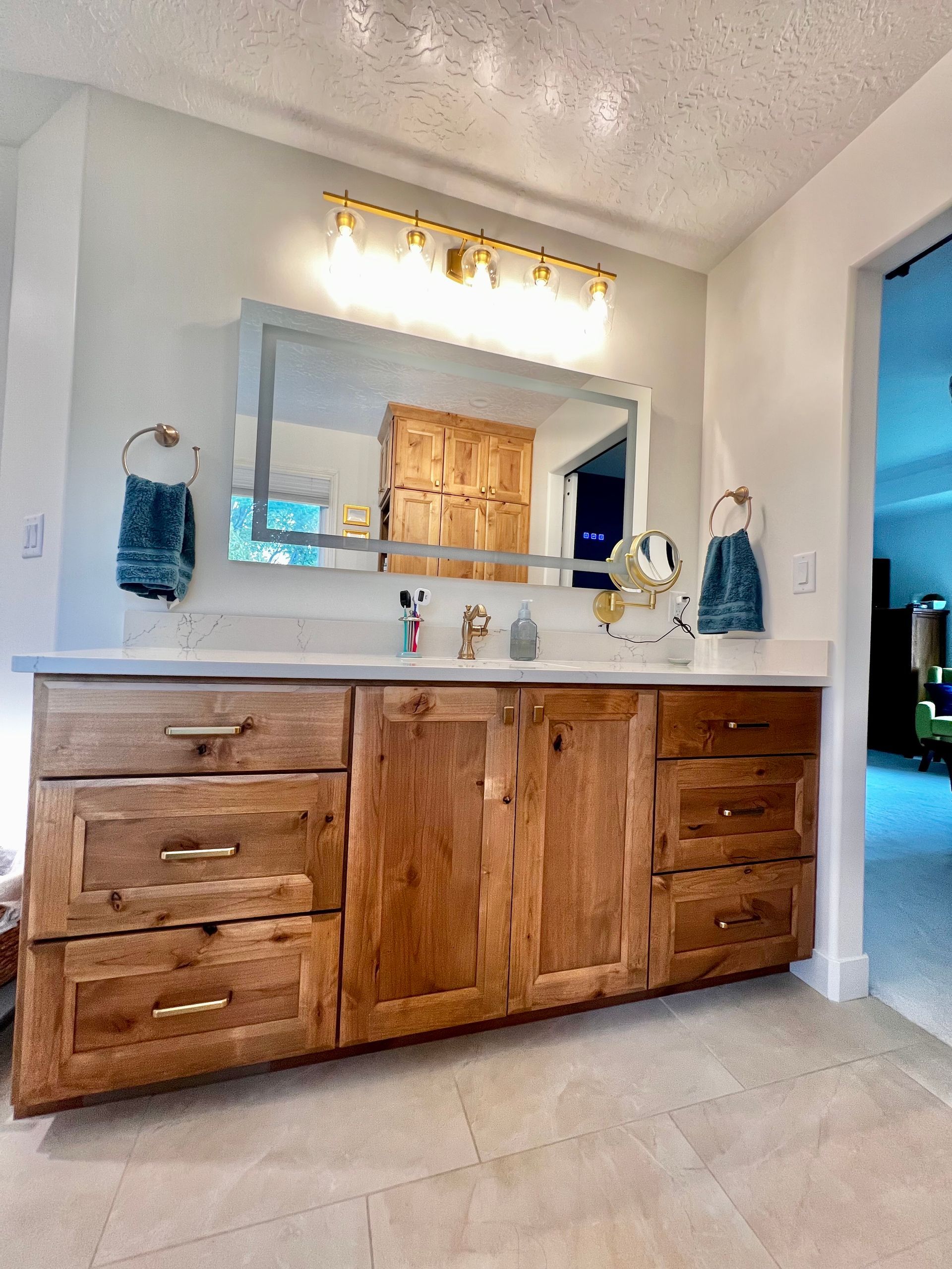 Bathroom vanity with natural wood cabinets, brass hardware, a wide mirror, and a bright light fixture.