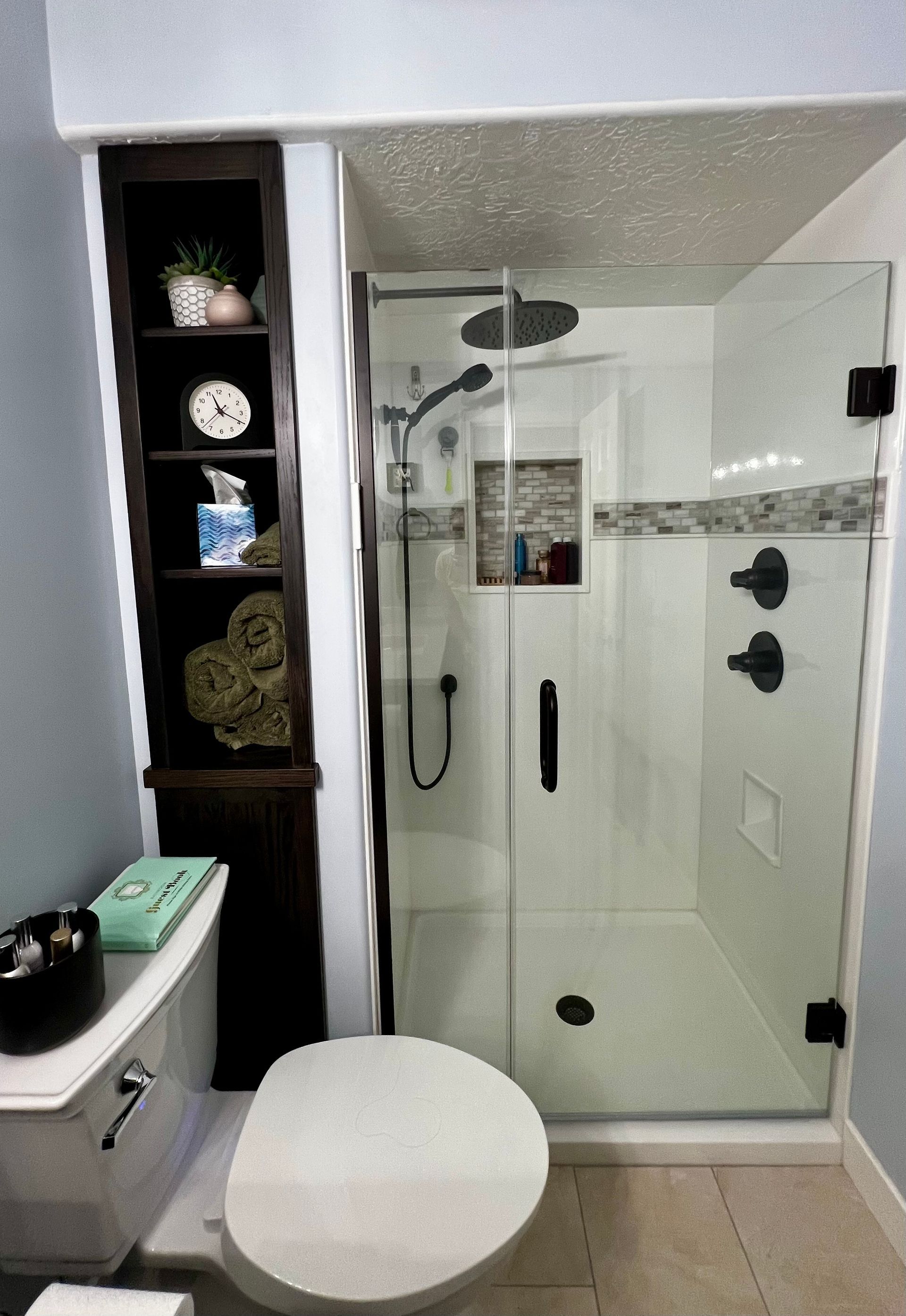 A bathroom featuring a glass shower stall next to a tall, dark wood shelving unit and a white toilet.