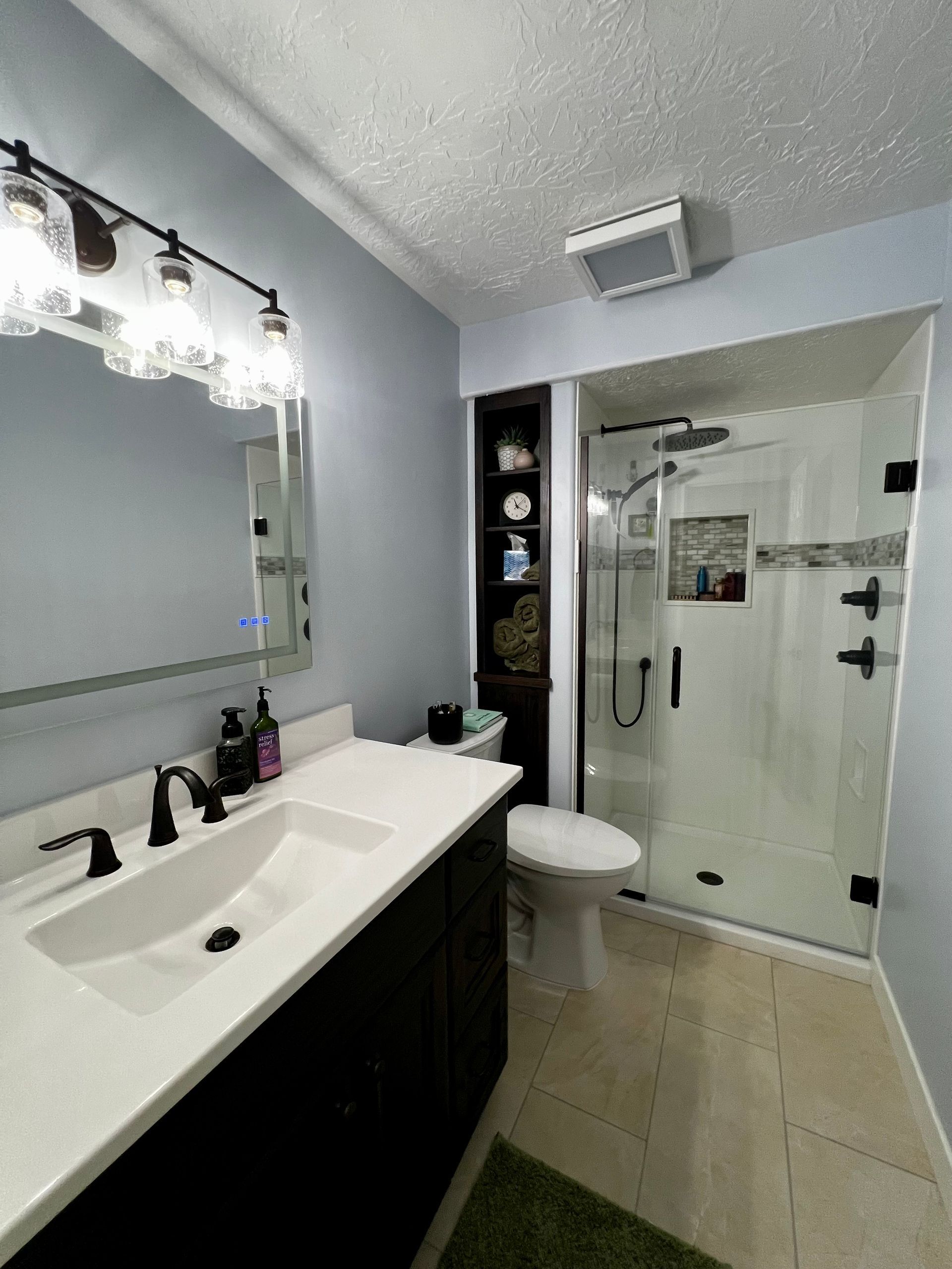 A modern bathroom with a black vanity, white countertop, illuminated mirror, dark shelving, and a glass-enclosed shower.