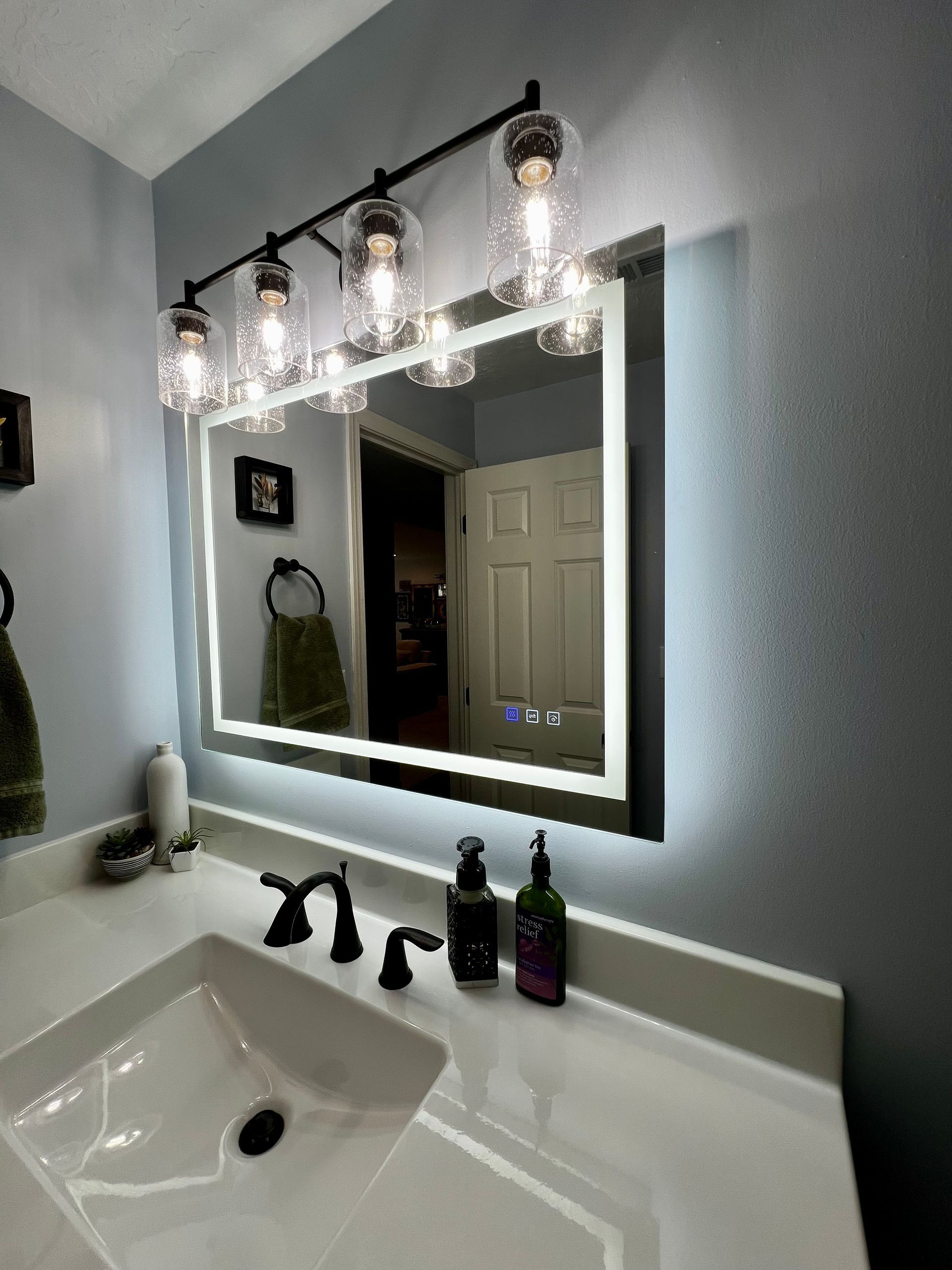 A bathroom vanity featuring a lighted mirror with a digital display, a four-bulb industrial light fixture, and a white sink.