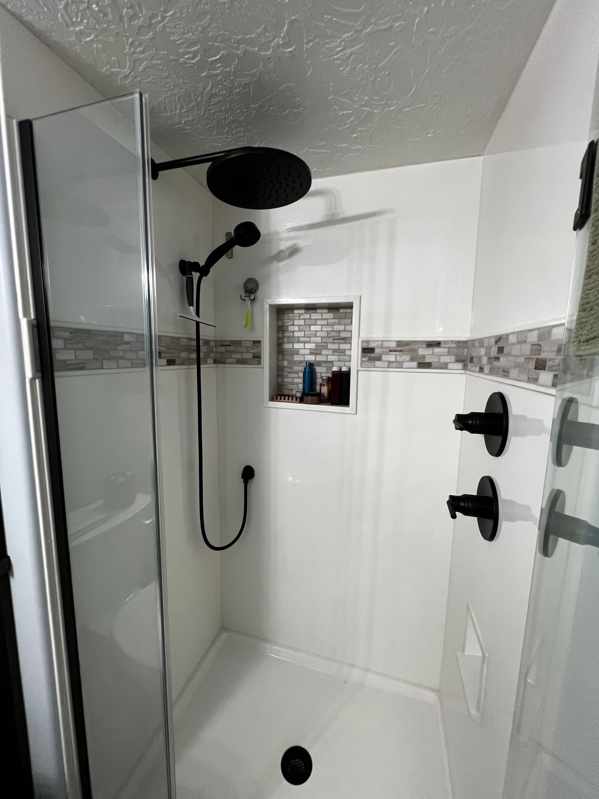 A walk-in shower with white tiled walls, a black rainfall showerhead, a handheld sprayer, and a built-in storage niche.