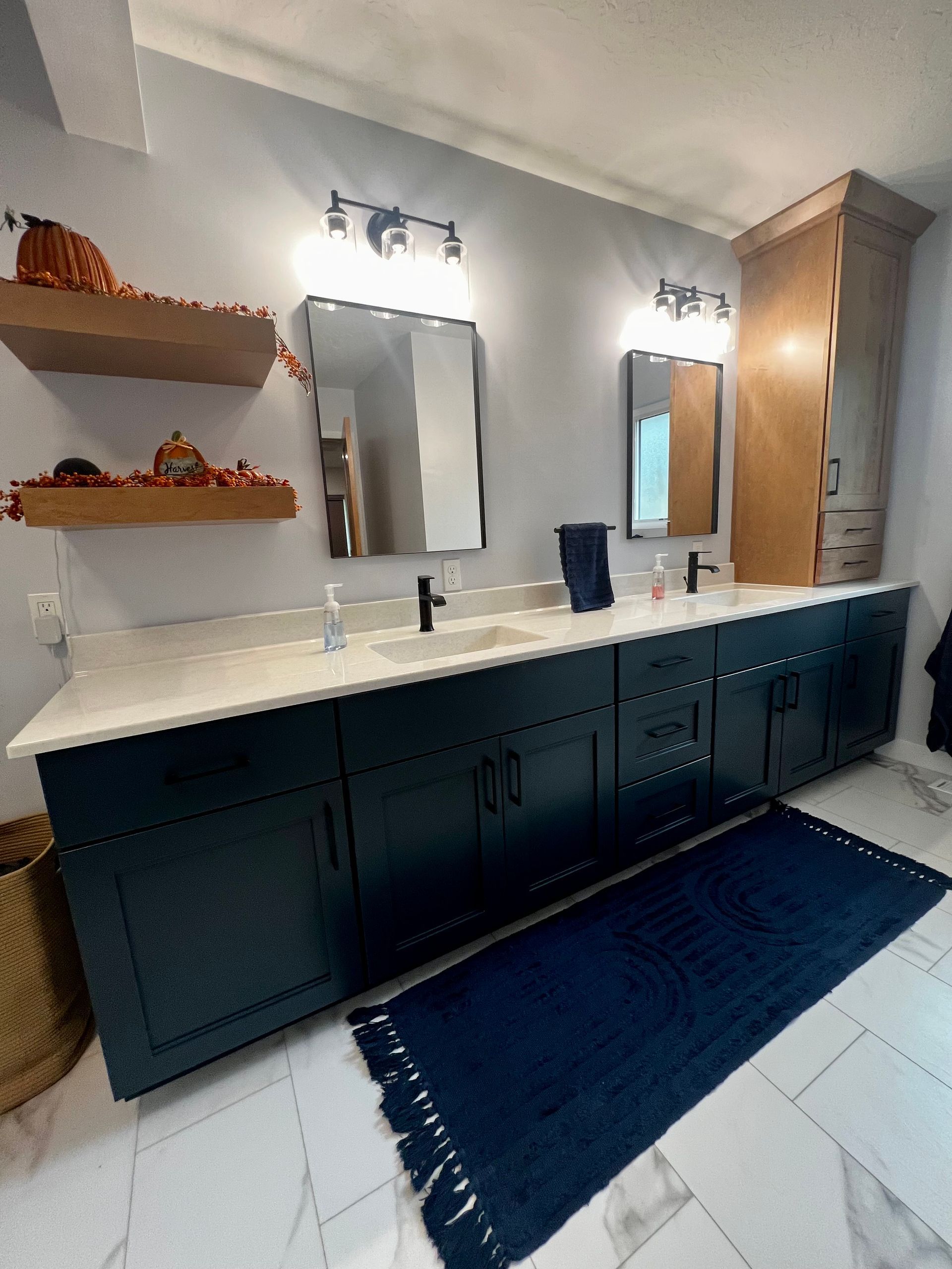A modern double vanity bathroom with dark cabinets, white countertops, two mirrors, and a blue rug on a marble tile floor.
