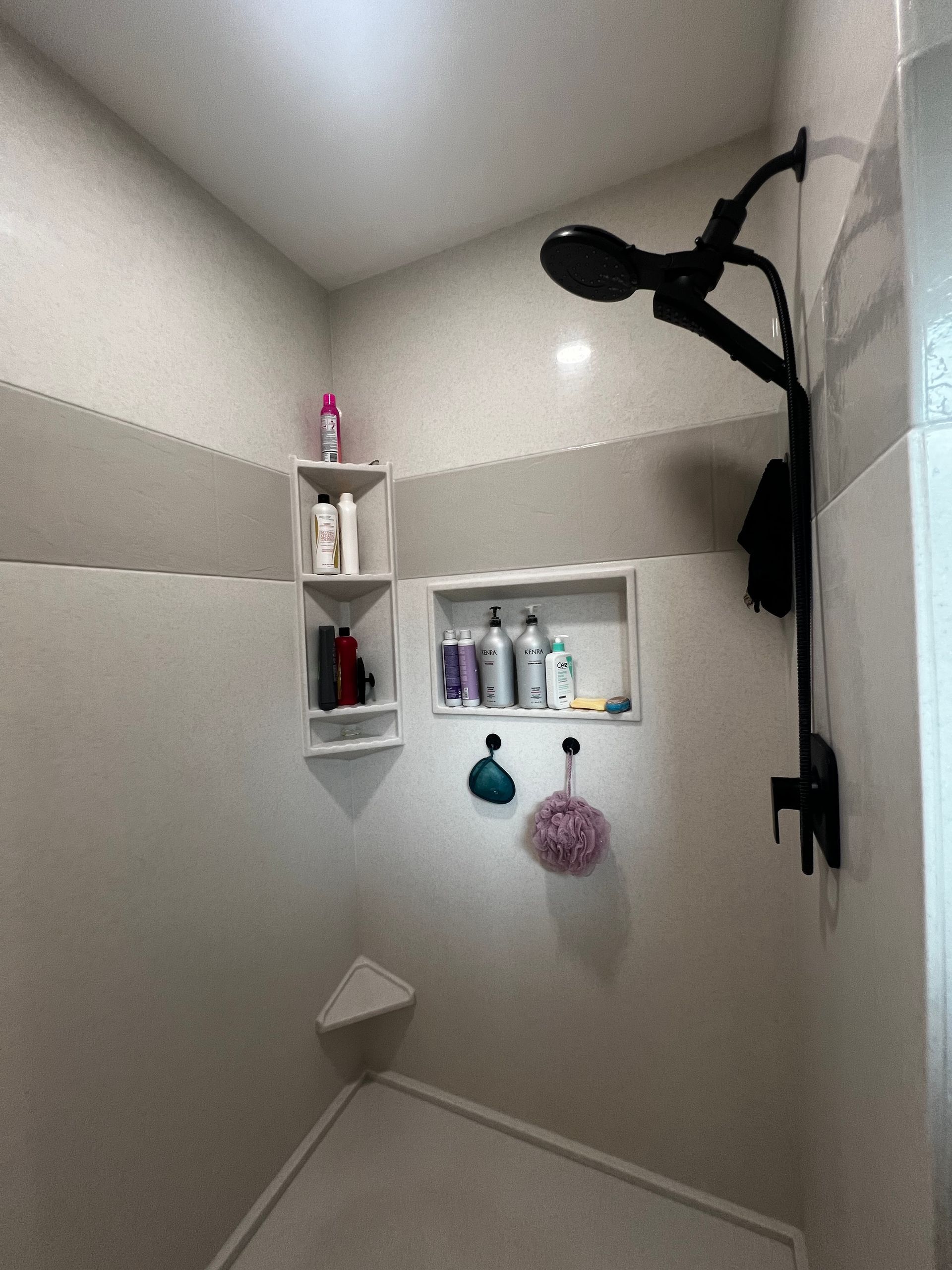 A walk-in shower with light-colored tile, black fixtures, a corner shelf, a built-in recessed niche, and hanging loofahs.