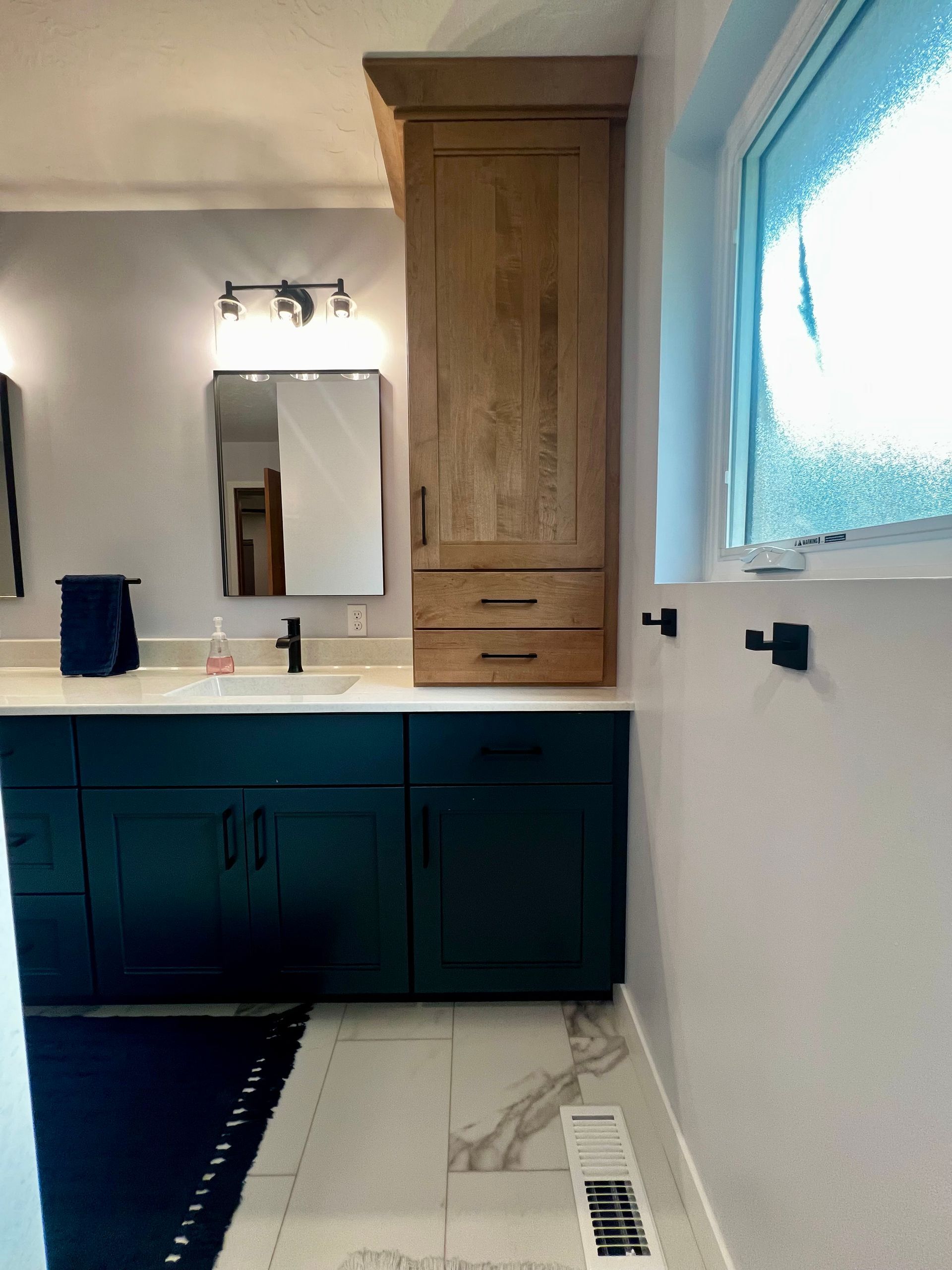 A bathroom vanity with dark teal cabinets, a tall wooden storage tower, white countertops, and black hardware.