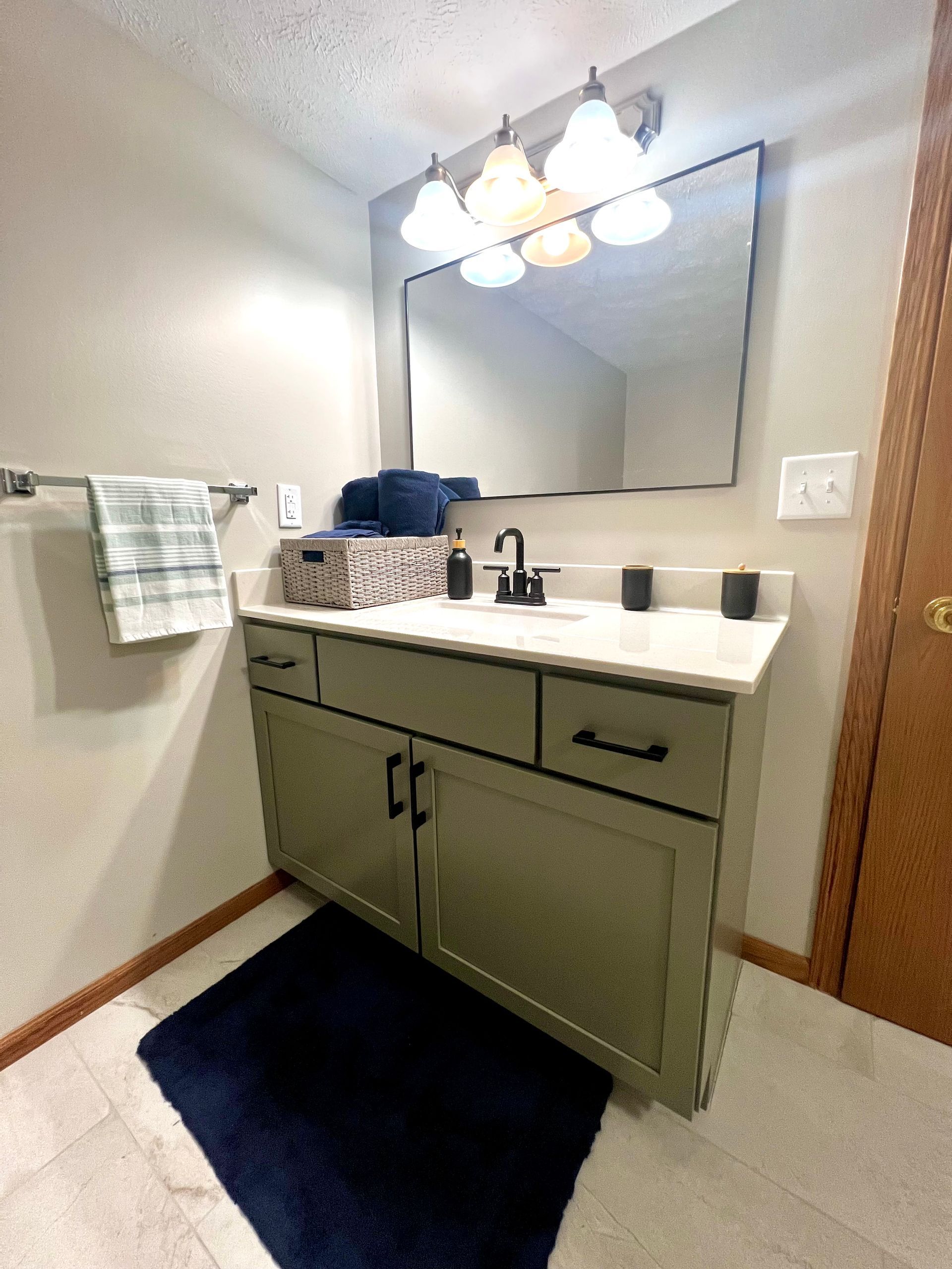 A bathroom vanity with olive-green cabinets, a white countertop, dark hardware, and a mirror under a three-bulb light fixture.