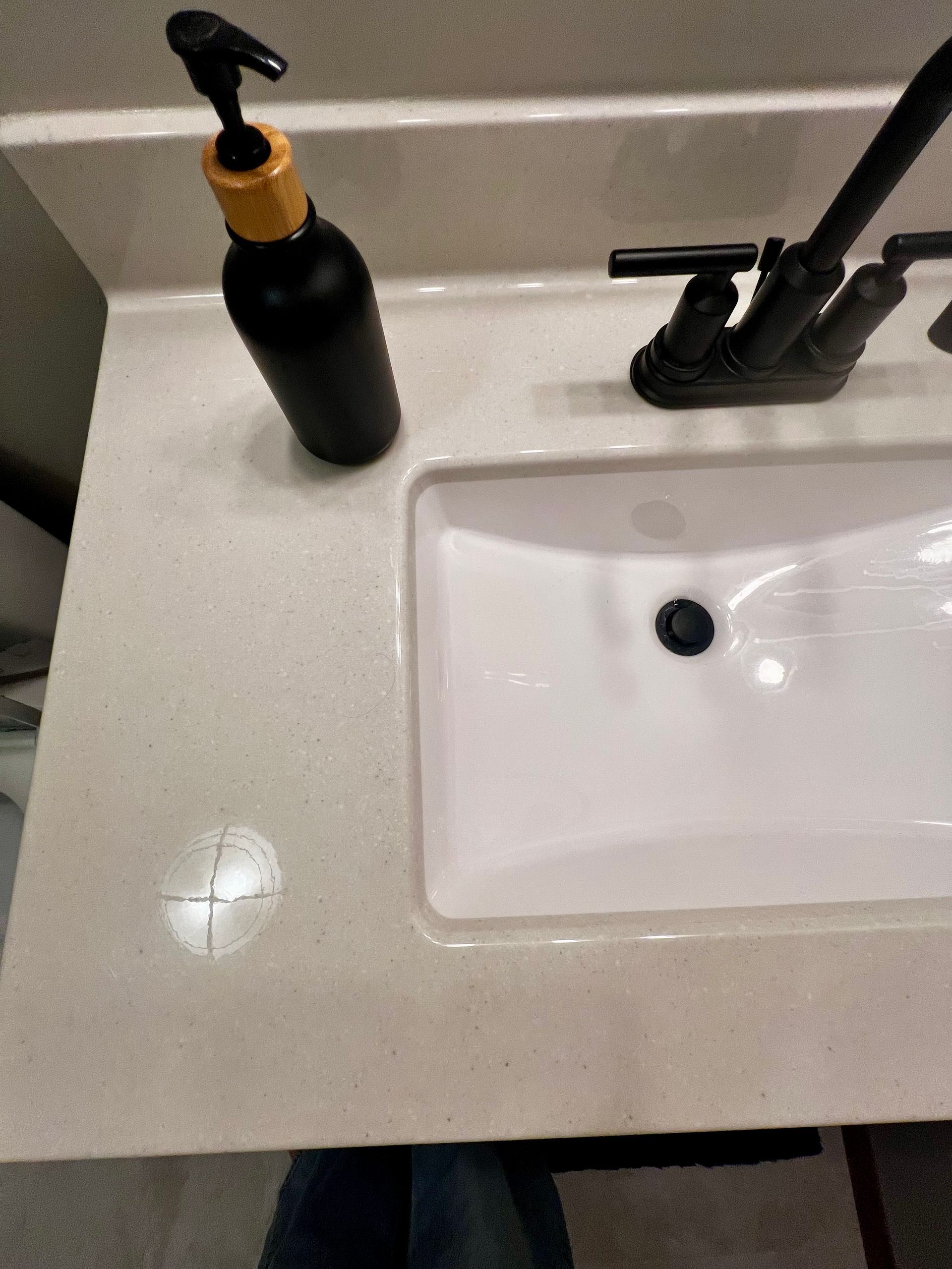 A black soap dispenser with a wooden pump rests on a light-colored speckled countertop next to a white sink basin.