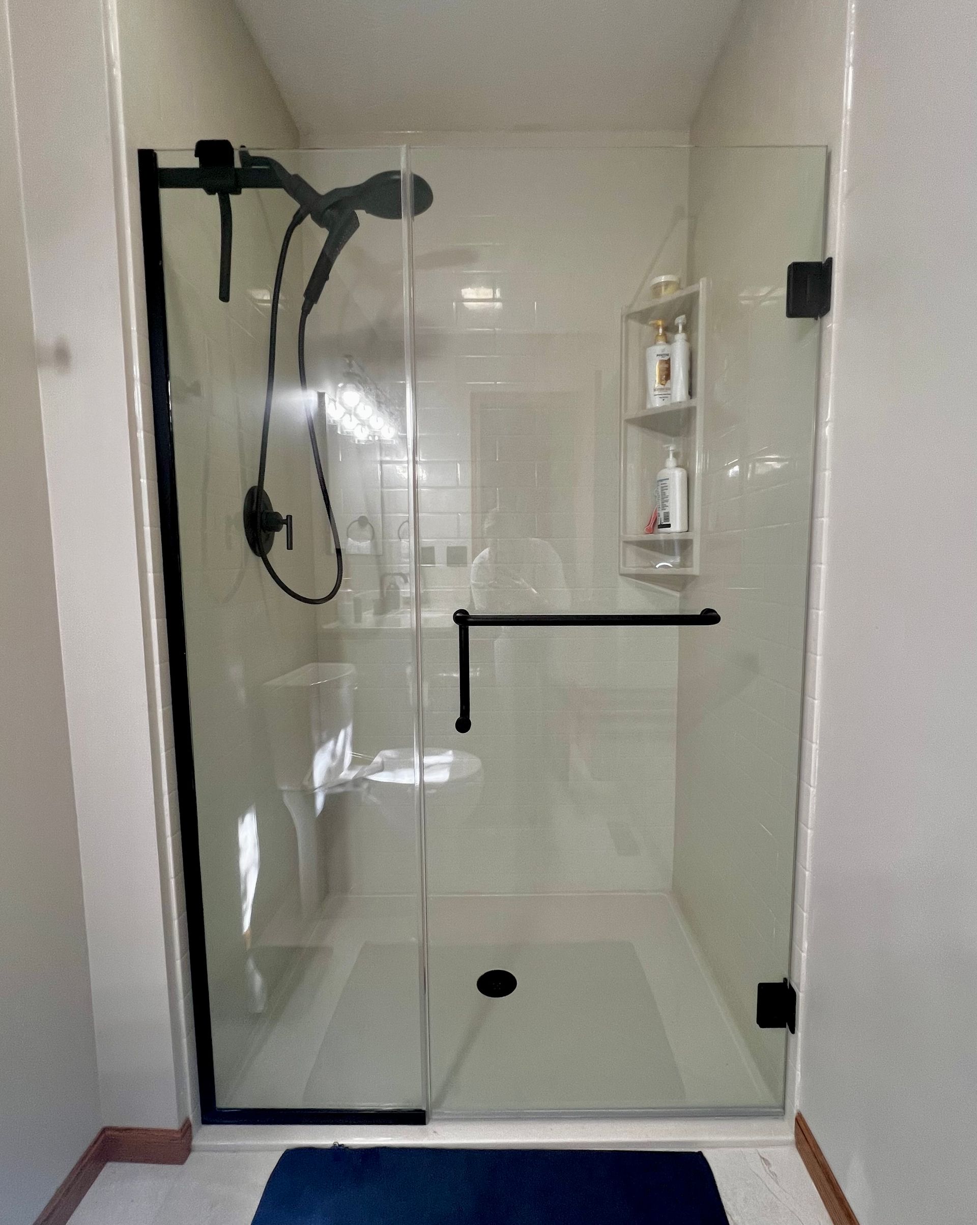A frameless glass shower door with matte black hardware and a handheld shower head inside a neutral-toned bathroom.