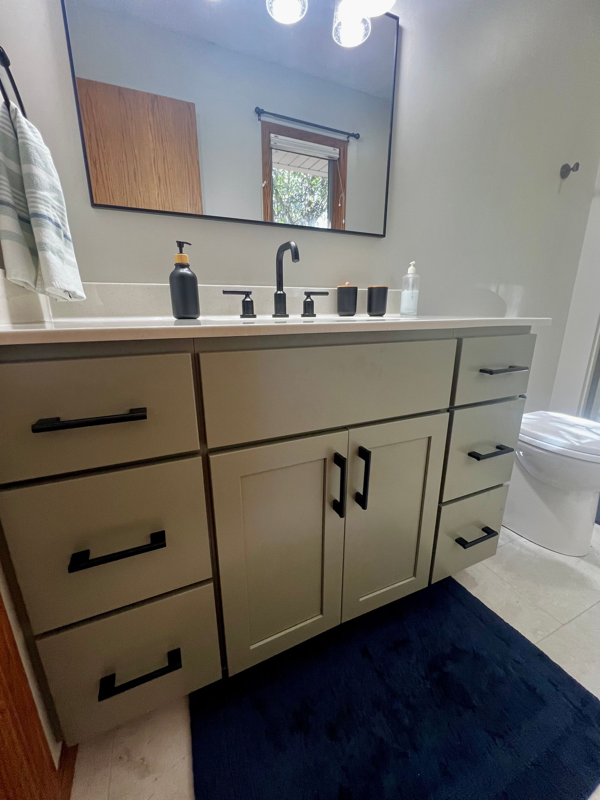 A bathroom vanity with light-colored cabinets, black hardware, a white countertop, sink, and mirror, over a blue rug.