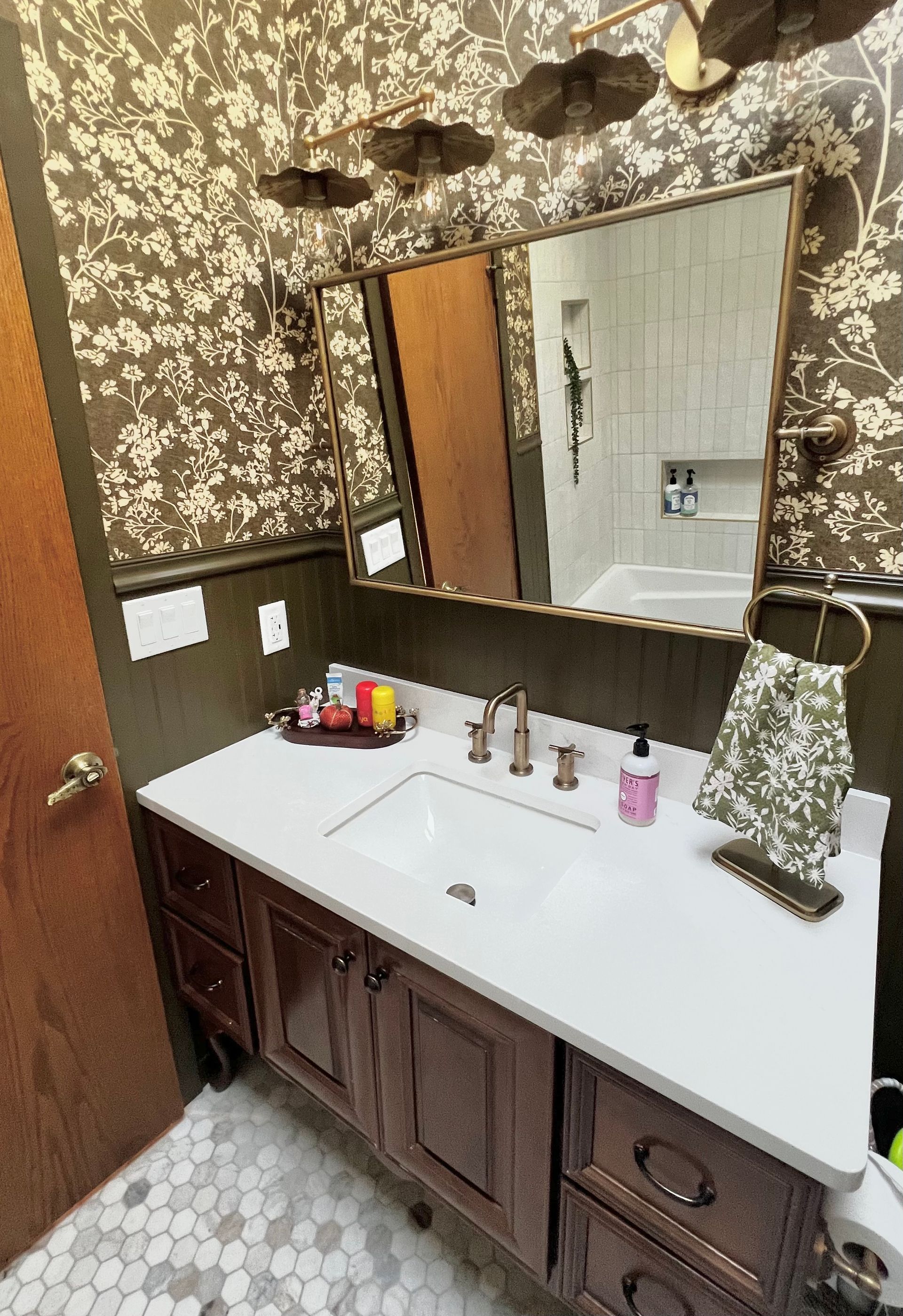 A bathroom vanity with a white countertop, dark wood cabinets, a mirror, and brown floral wallpaper.